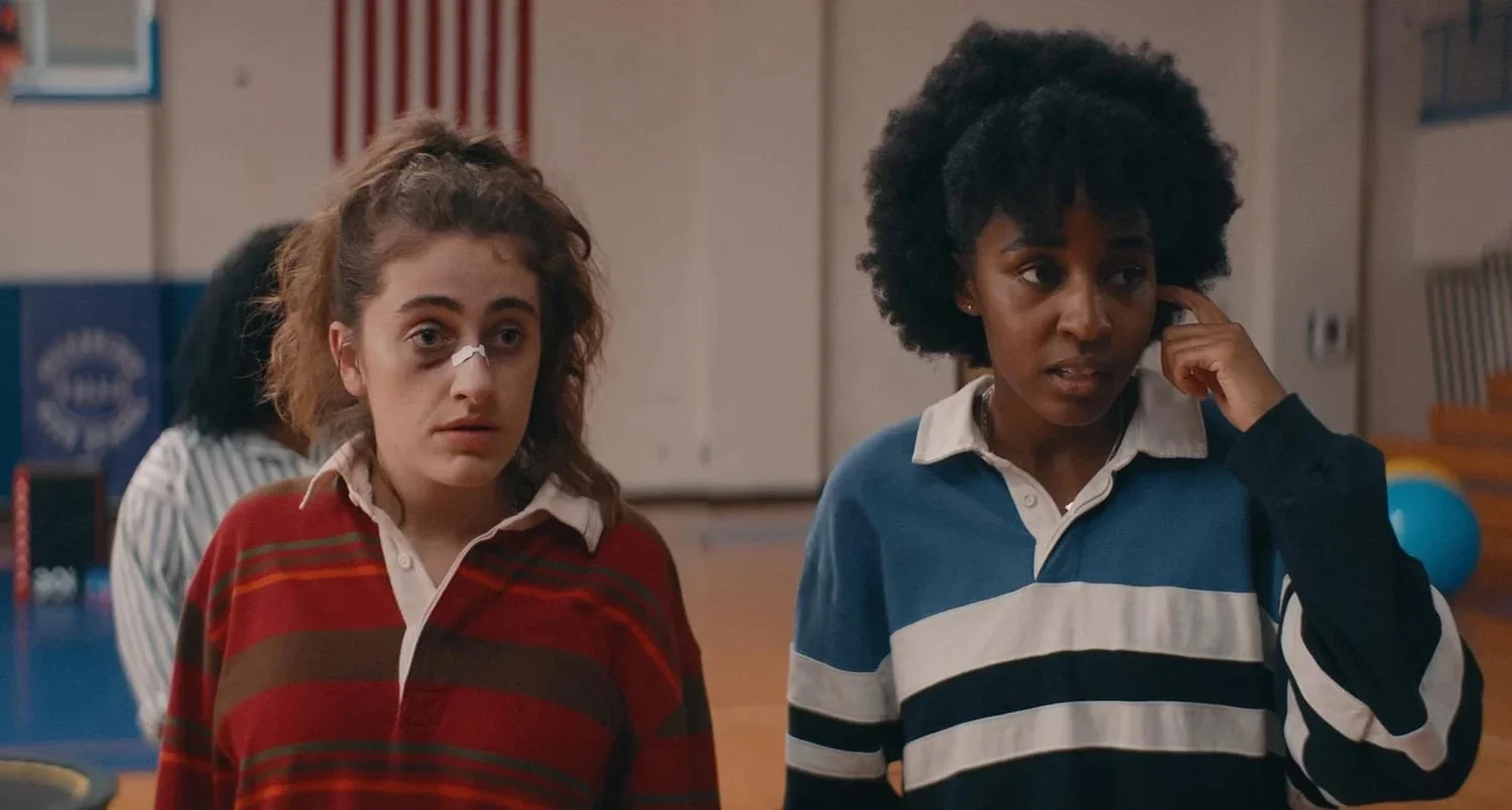 two girls staring off camera. One has a broken nose in a red striped shirt. The other has a blue striped polo shirt.