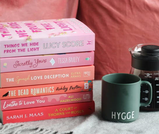 A stack of pink and red romance and romantasy books, with a green coffee mug and coffee pitcher next to it