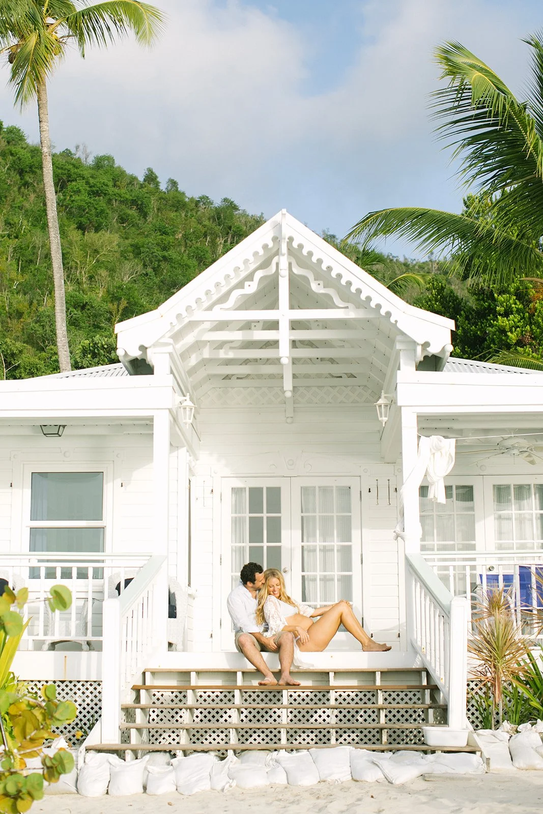 A couple sitting on the front steps of a white beach house, surrounded by sand and tropical plants, with hills and palm trees in the background.