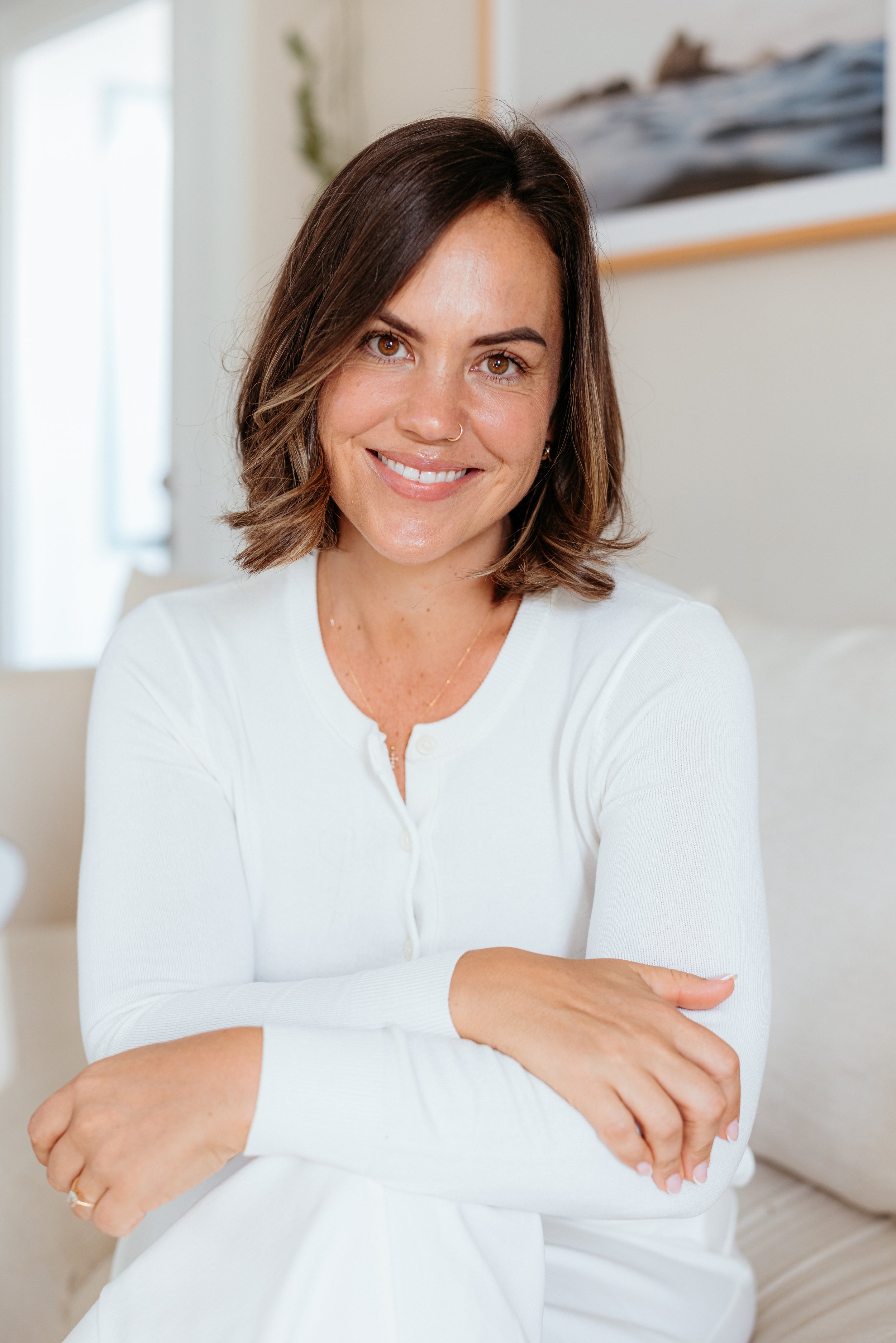 A woman with shoulder-length brown hair and brown eyes, smiling while sitting indoors in front of a neutral background with artwork on the wall.