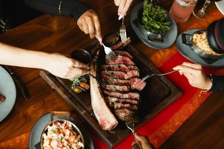 Top-down view of sliced steak on a wooden board as multiple hands use forks to serve, surrounded by plated side dishes.