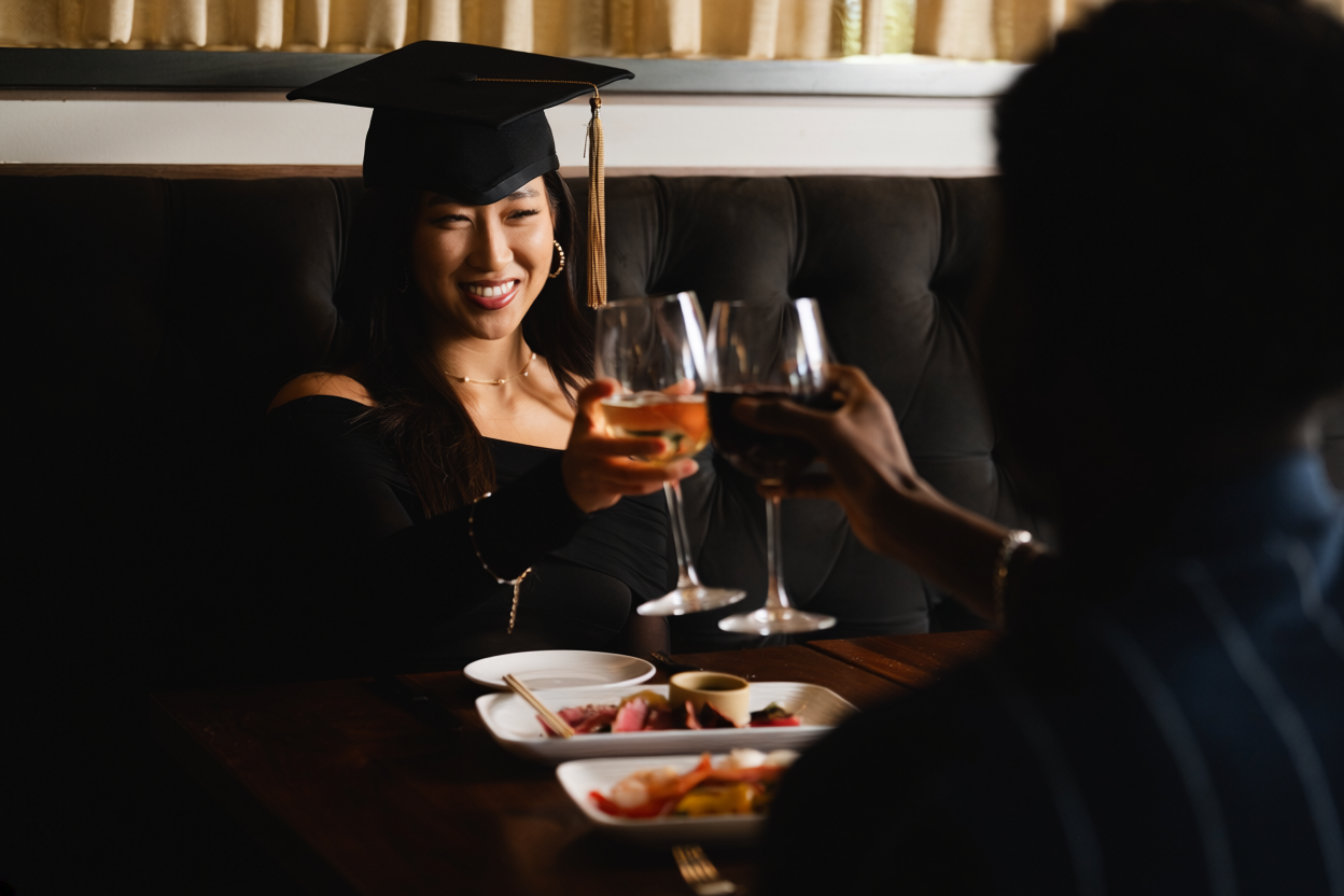 wo adults seated at a restaurant table, each wearing a black graduation cap, raising glasses over plated food in a dim indoor setting.
