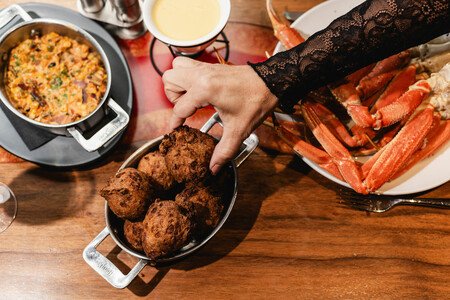 Overhead view of a dining table with a hand serving hushpuppies, crab legs on a plate, and seafood dishes arranged on a wooden table.