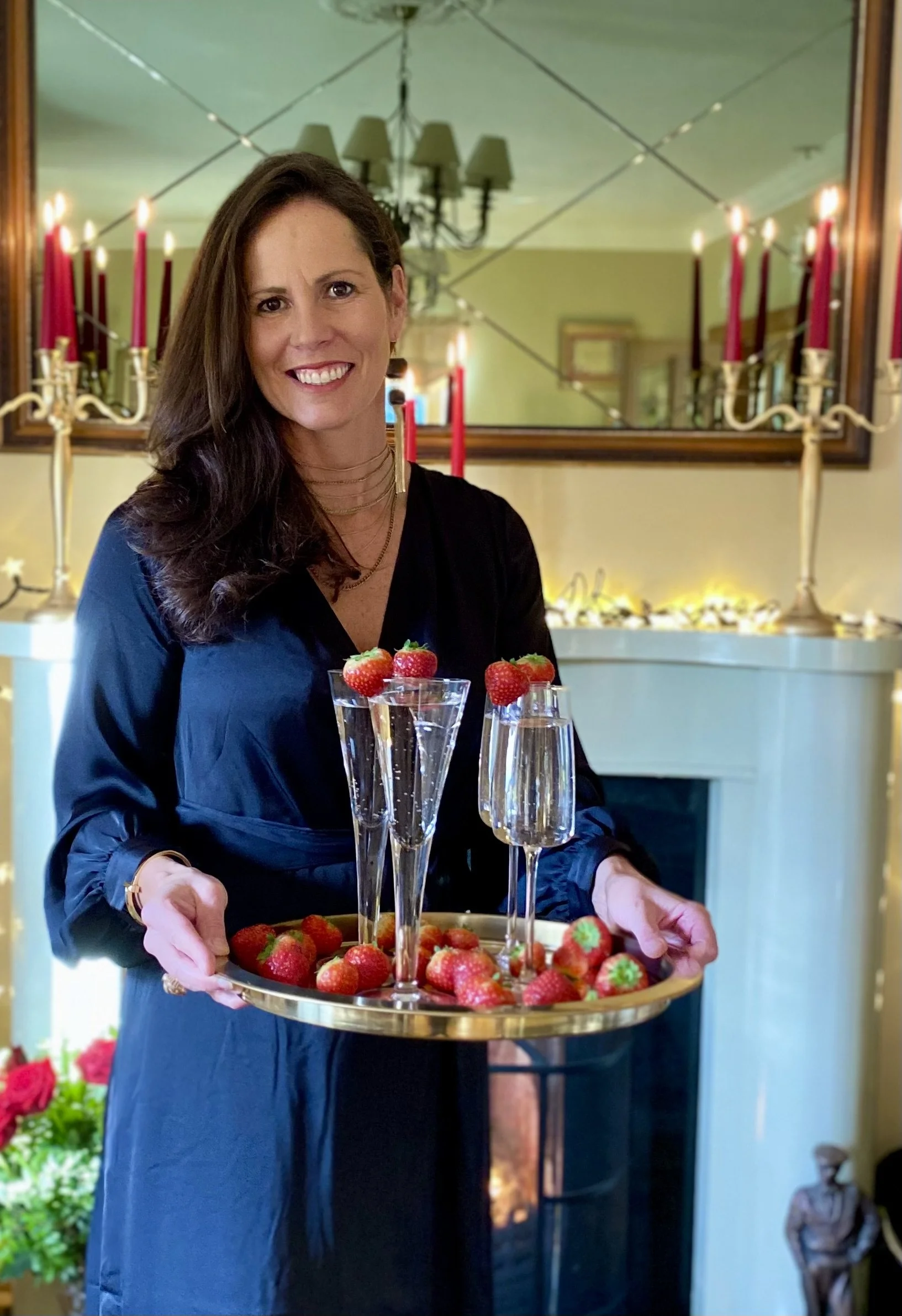 A woman in a black dress holding a tray with glasses of champagne and strawberries in a decorated living room.
