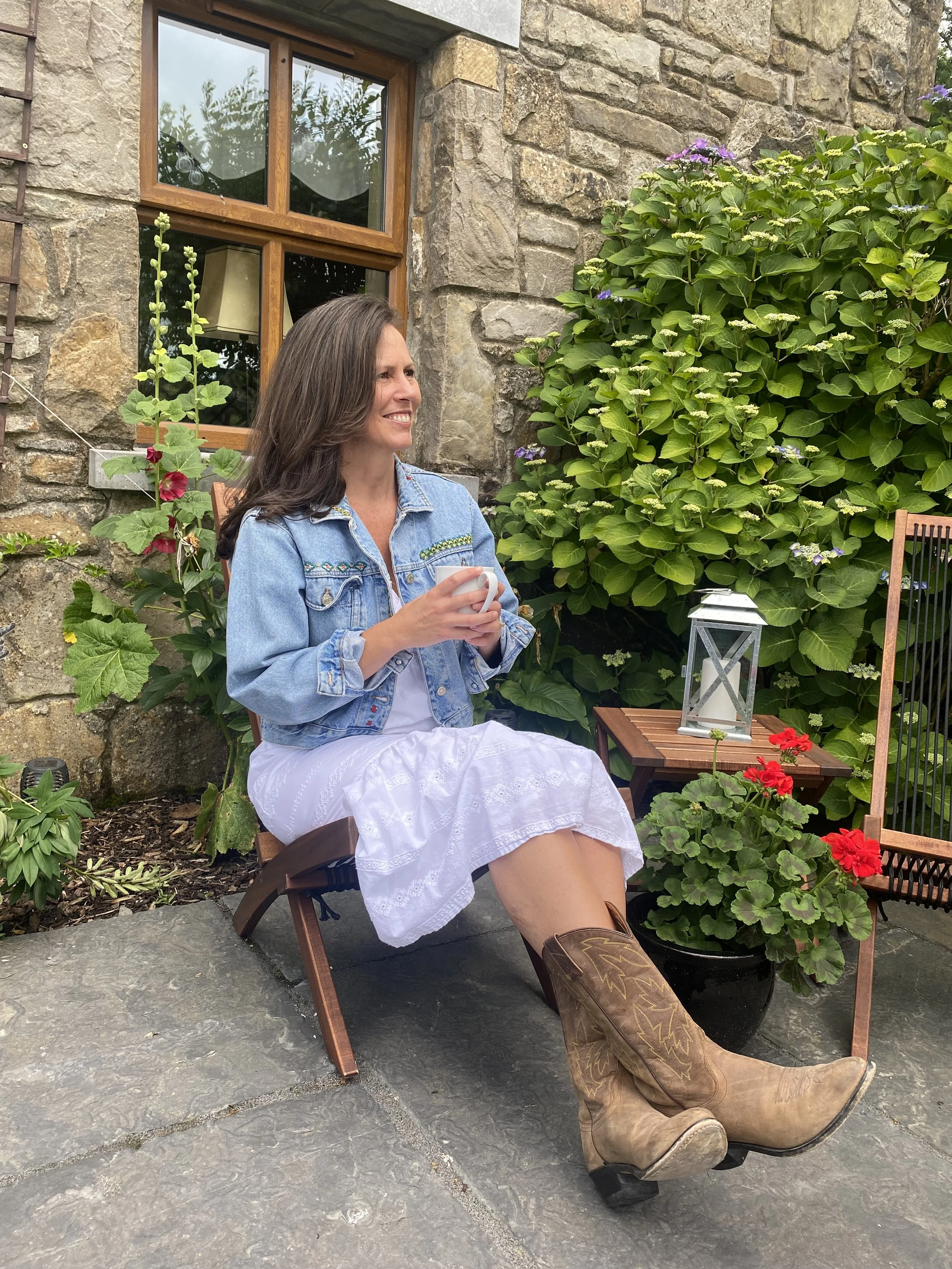 A woman sitting outdoors on a foldable chair, holding a mug, smiling, in front of a stone wall and lush green bushes with purple and red flowers. She is dressed in a white dress, a blue denim jacket, and brown cowboy boots.