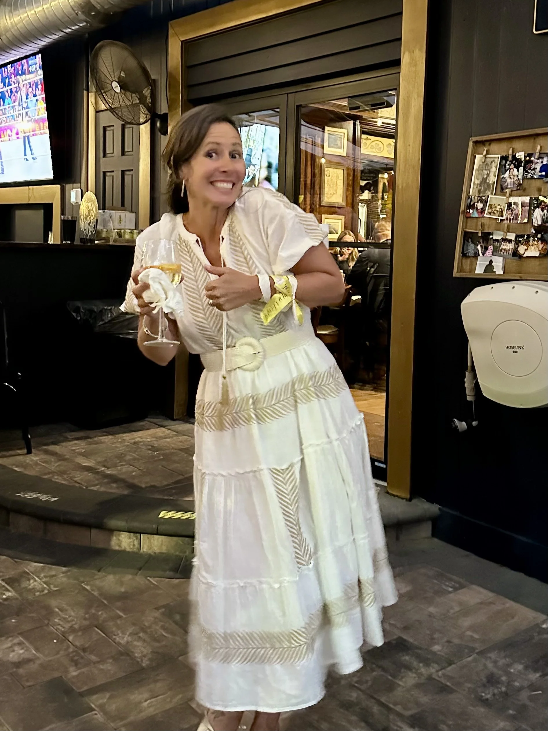 A woman in a white dress with gold leaf patterns holding a glass of wine, standing in a restaurant with a rustic interior.