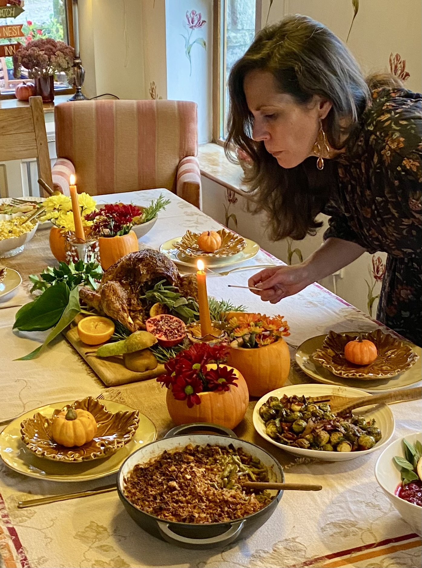 Woman decorating a Thanksgiving dinner table with pumpkins, candles, and various dishes, including a turkey, Brussels sprouts, and sweet potatoes.