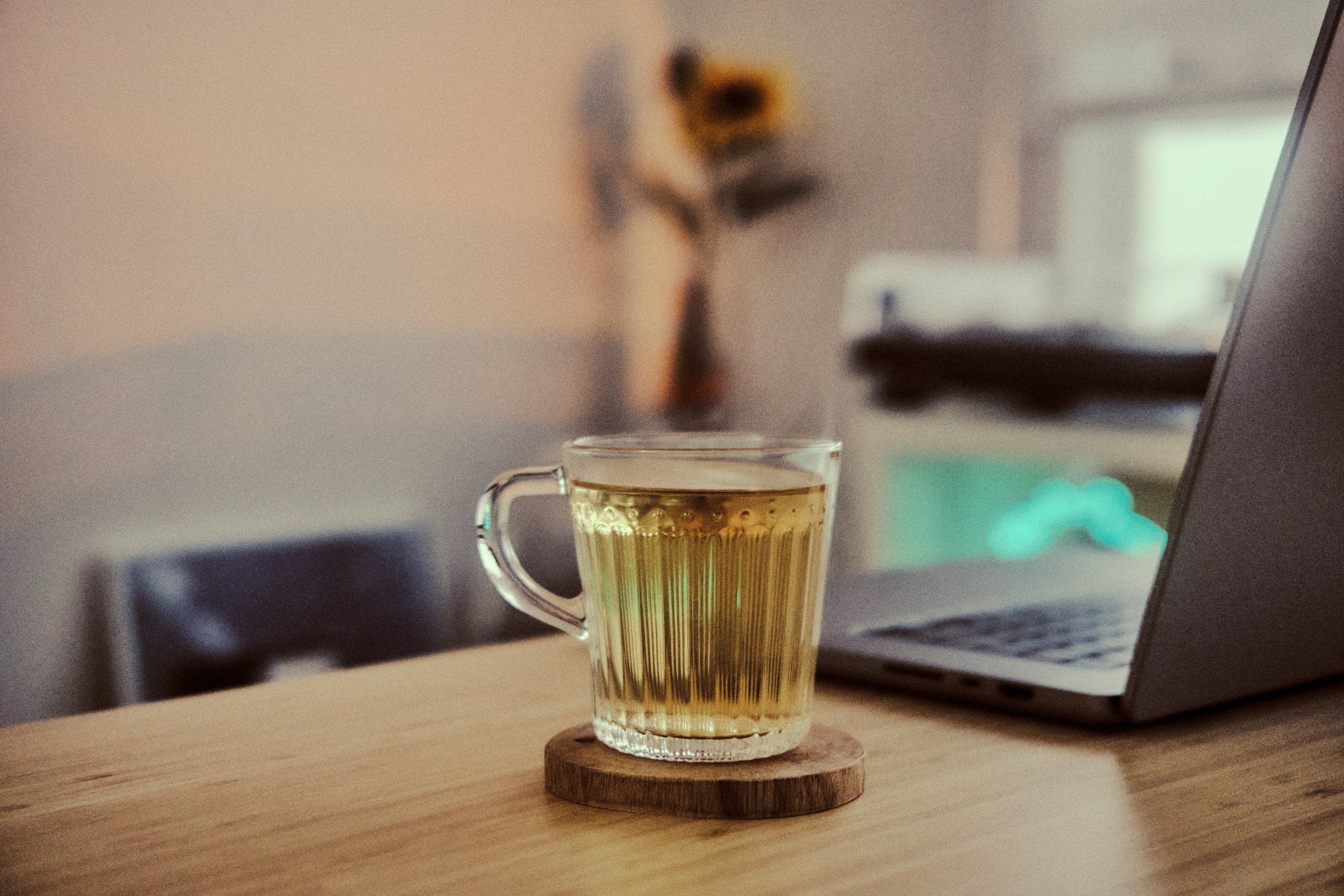 A clear glass cup filled with a hot beverage, placed on a small round wooden coaster on a light-colored wooden surface, with a laptop and blurred background.