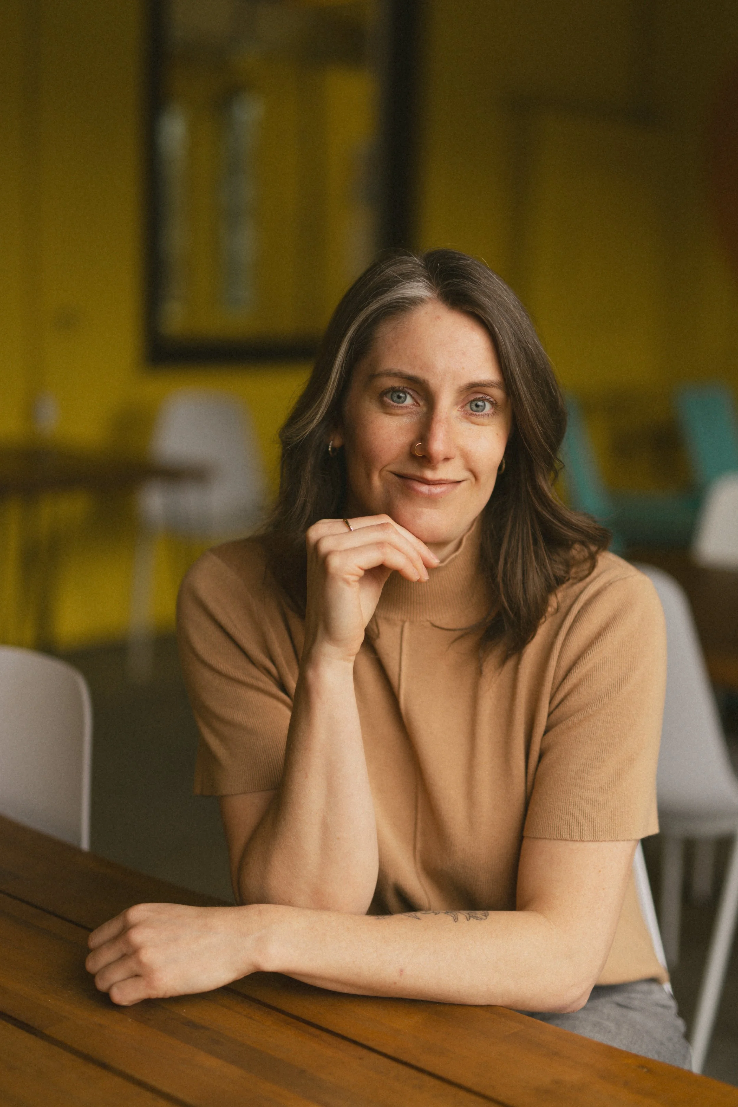 A person with brown hair, blue eyes, and a nose ring wearing a brown shirt, sitting at a wooden table in a room with yellow walls, smiling at the camera, with her chin resting on their hand.