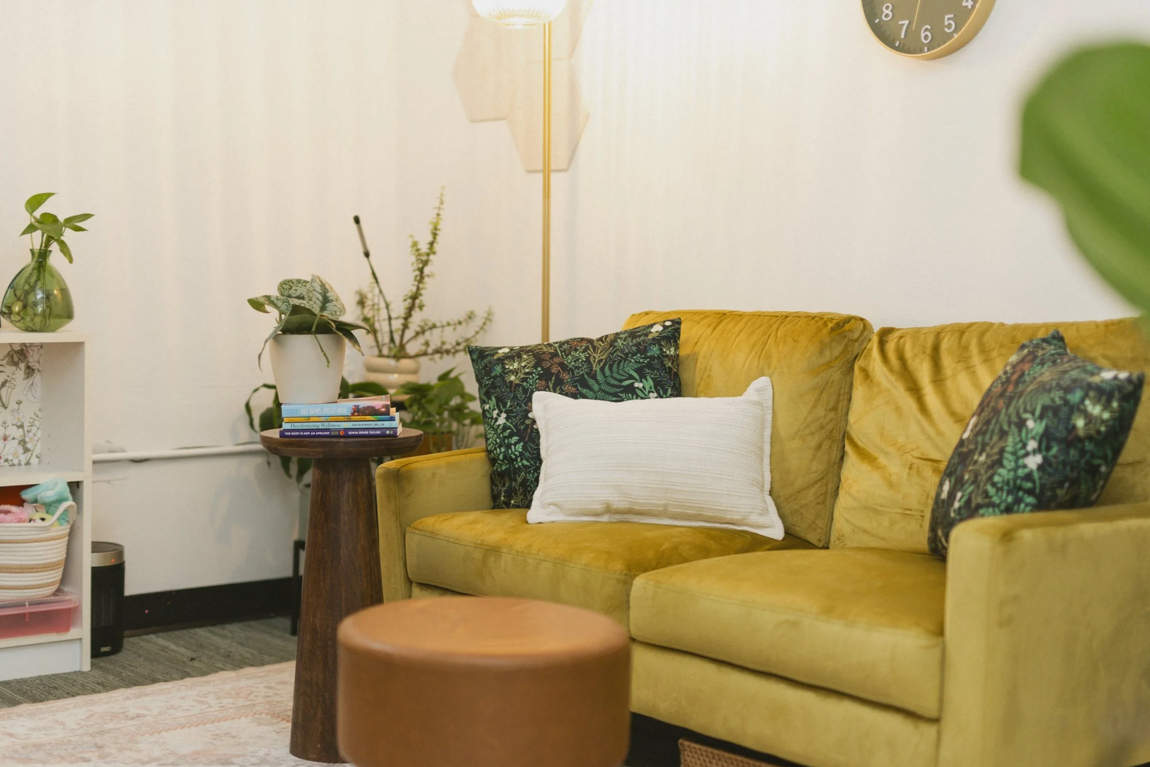 Living room with a yellow sofa, decorative pillows, a small wooden side table with books, potted plants, a wall clock, a floor lamp, and shelves with fidget items.