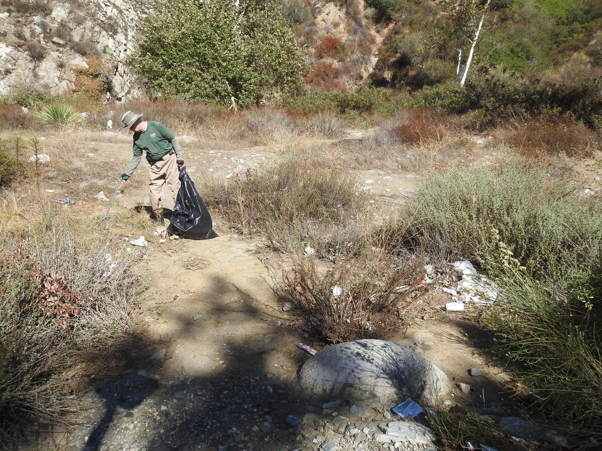 FRVC Volunteer picking up trash in a desert-like natural area with bushes, rocks, and dry vegetation.