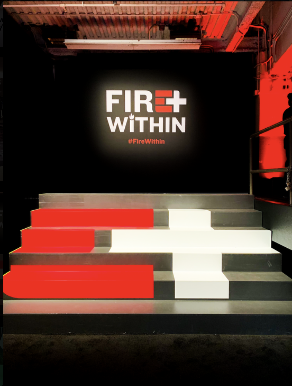 A black wall with a logo that says 'FIRE WITHIN' in bold white letters, with an illuminated background and a red hashtag '#FireWithin' underneath. There are steps in front with red, white, and gray color blocks, and red lighting highlighting the envi
