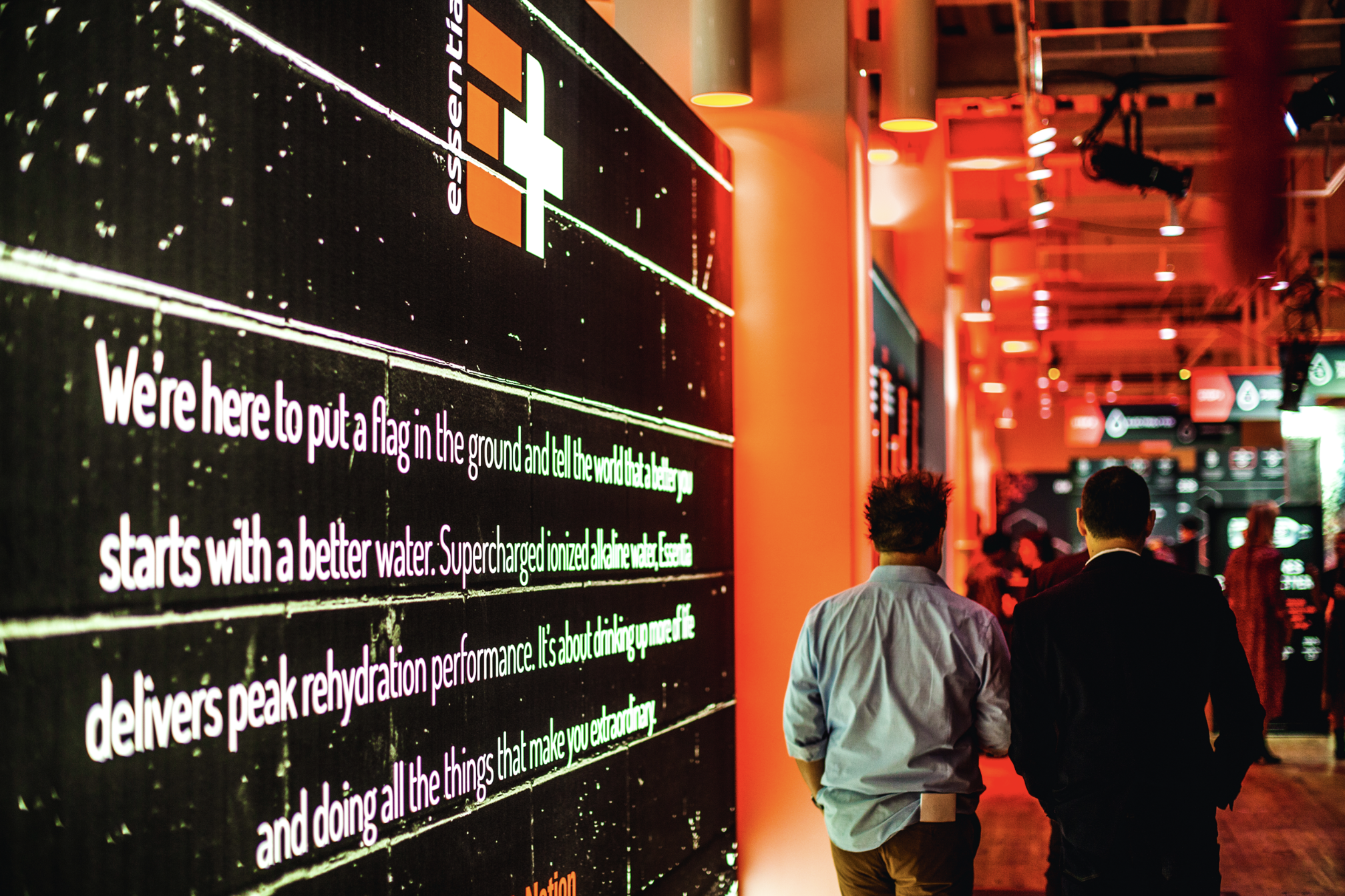 Two people walking down a brightly lit hallway next to a large digital screen displaying the Esphage logo and text about hydration.