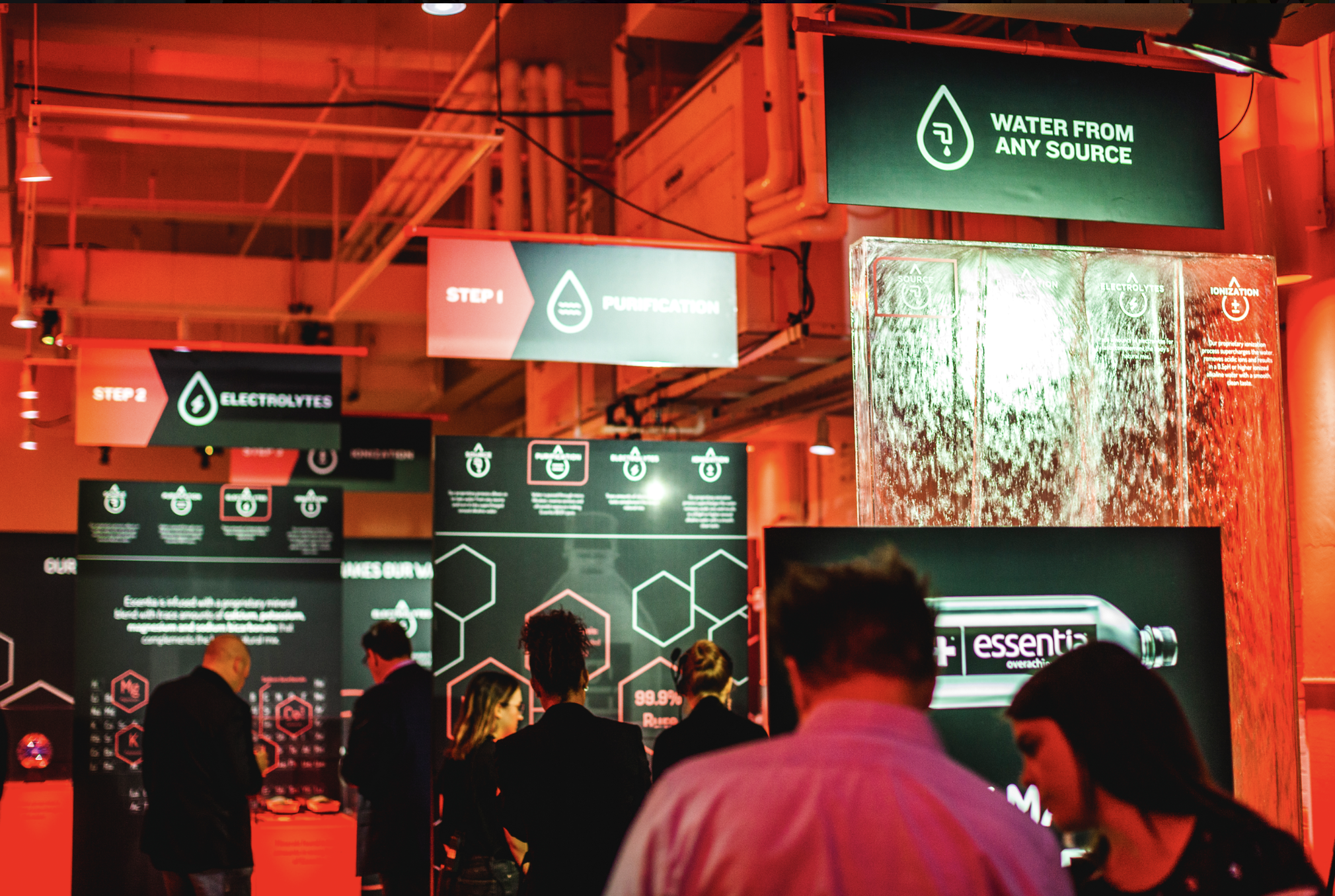 Indoor exhibition with large digital screens displaying information about water purification, electrolytes, and ionization. Several people are observing the displays, and the environment is illuminated with red and green lighting.