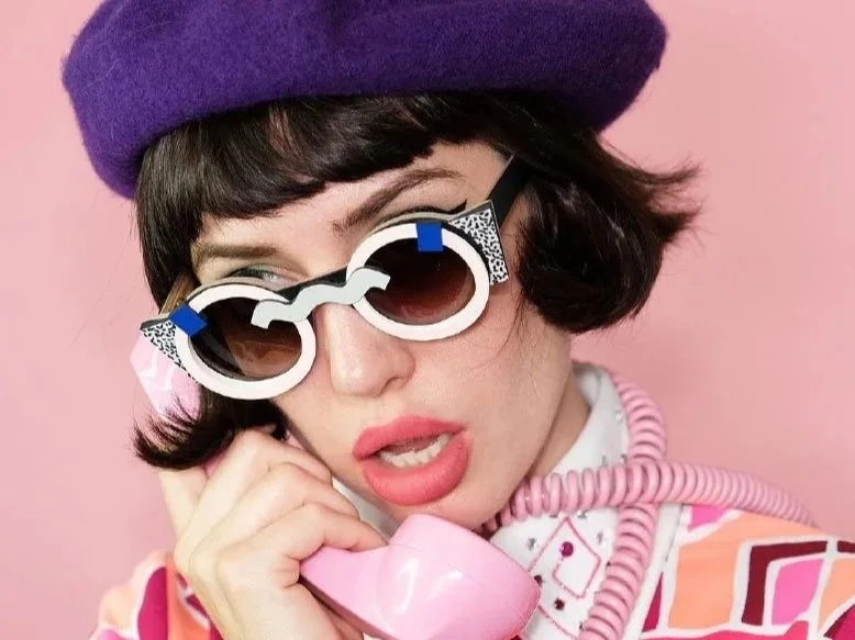 Woman wearing a purple beret and large white sunglasses holding a pink rotary phone.