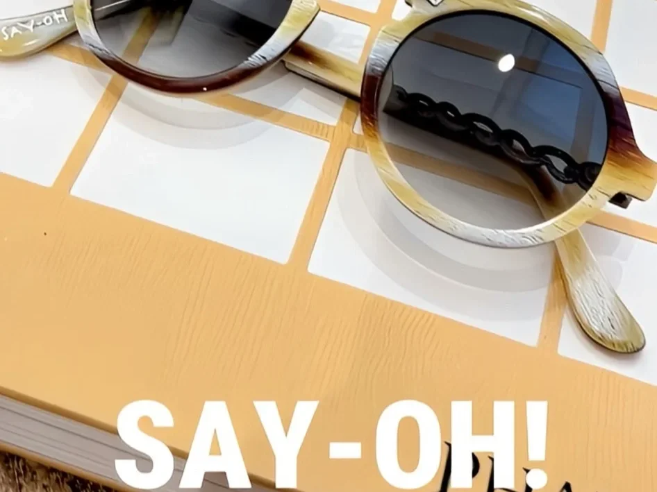 Two pairs of sunglasses, one with a tortoiseshell frame and the other with a cream and brown frame, are resting on a tiled surface. The foreground sunglasses have round, dark lenses and decorative metal detailing on the temples. The background sunglasses have a more rectangular shape with brown frames. The surface they are on is a kitchen or dining table with a grid pattern, and a large orange book with white text reading "SAY-OH!" is partially visible at the bottom.