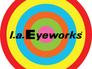 Close-up of a woman with bright red hair, wearing red cat-eye glasses and leopard print top. Bright, colorful background with yellow, purple, and blue abstract patterns. Text on image reads "l.a. Eyeworks - UNCENSORED VISIONS."
