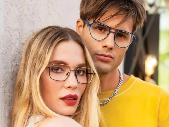 A young man and woman wearing glasses, leaning against a wall outdoors.