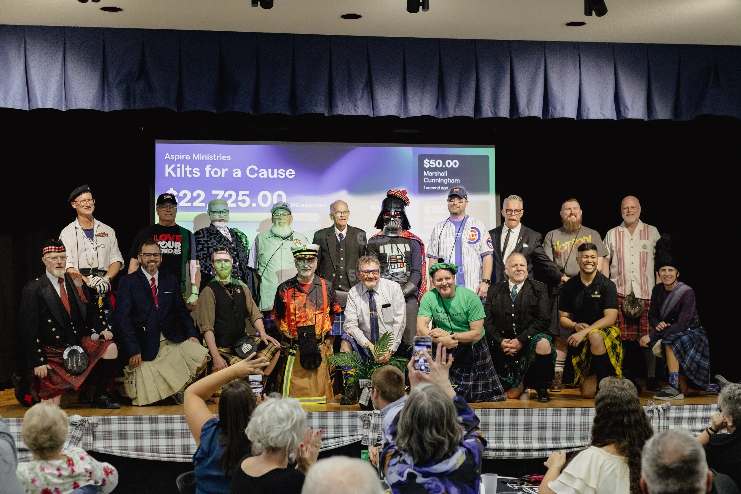 Image of 19 kilted men standing on stage for group photo