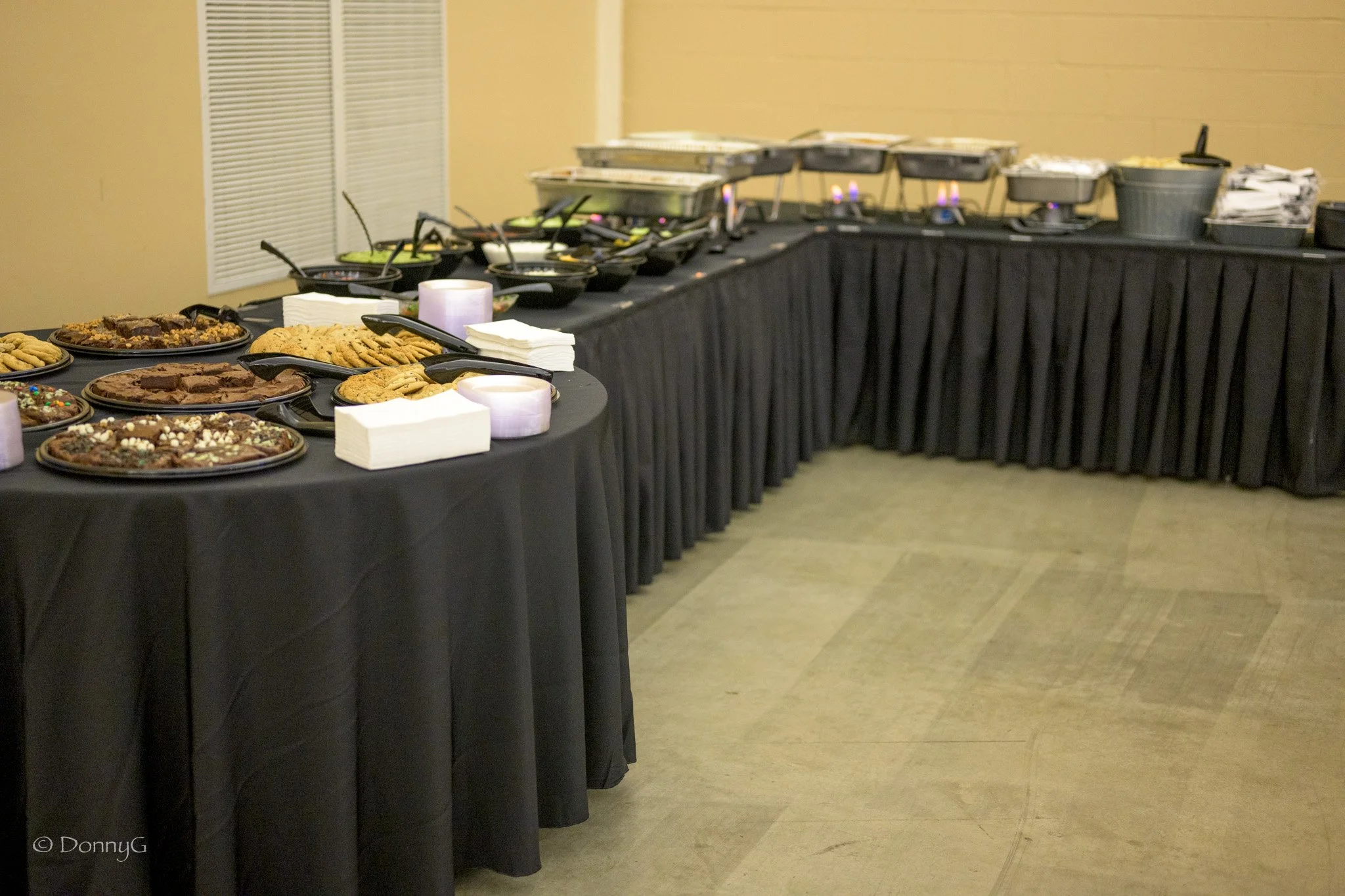 Buffet table with cookies, brownies, and snacks, covered with a black tablecloth, in an indoor setting.