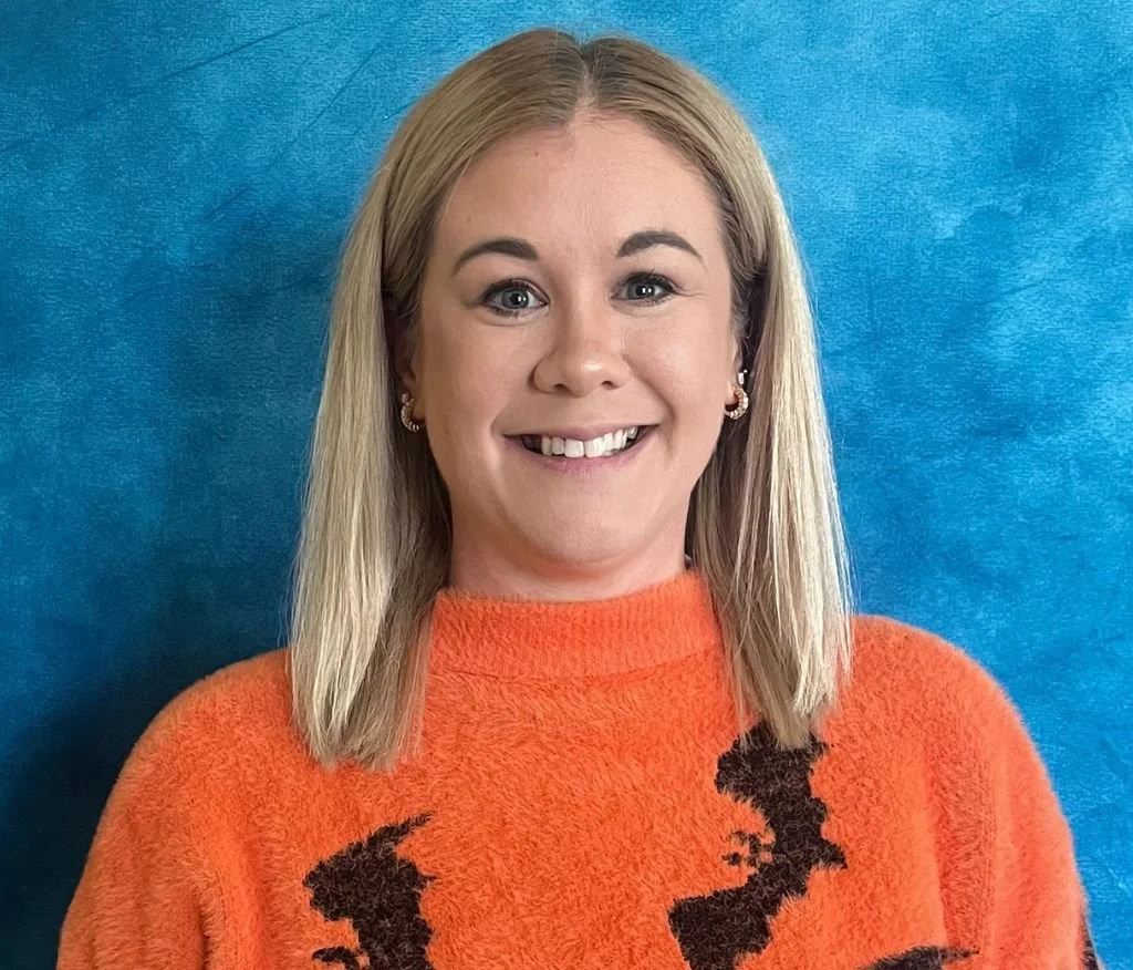 This is a picture of play therapist, Andrea Toledo, who offers play therapy in Schertz, TX. She is wearing an orange sweater and is standing in front of a blue background.
