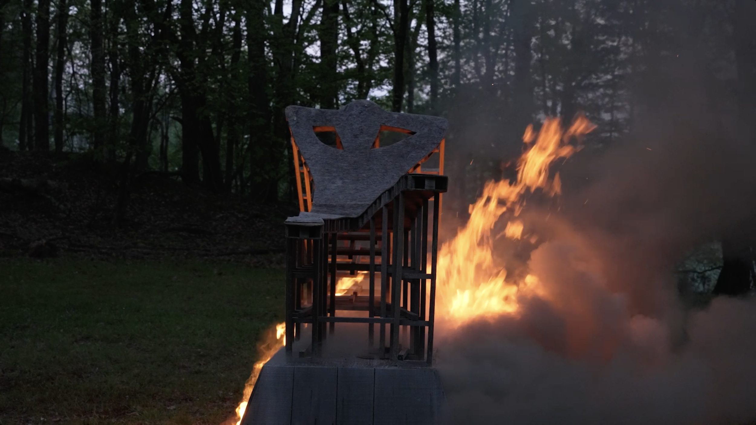 Outdoor scene at dusk with a wooden sculpture on a platform and a controlled fire burning nearby, surrounded by trees.