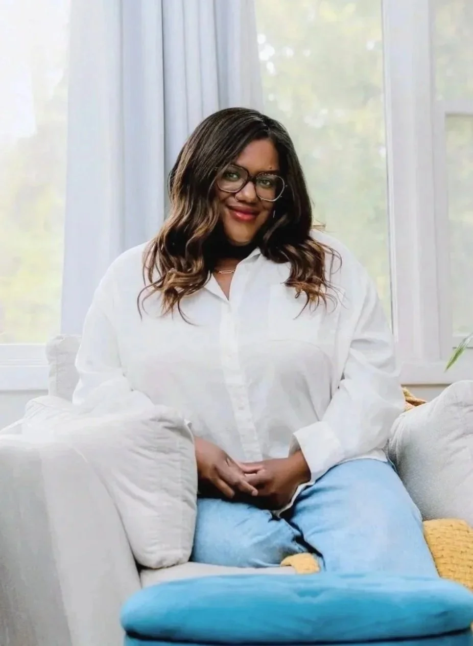 A woman with shoulder-length wavy hair wearing glasses, a beige wrap dress, and a cream-colored knit cardigan, smiling and looking at the camera indoors.