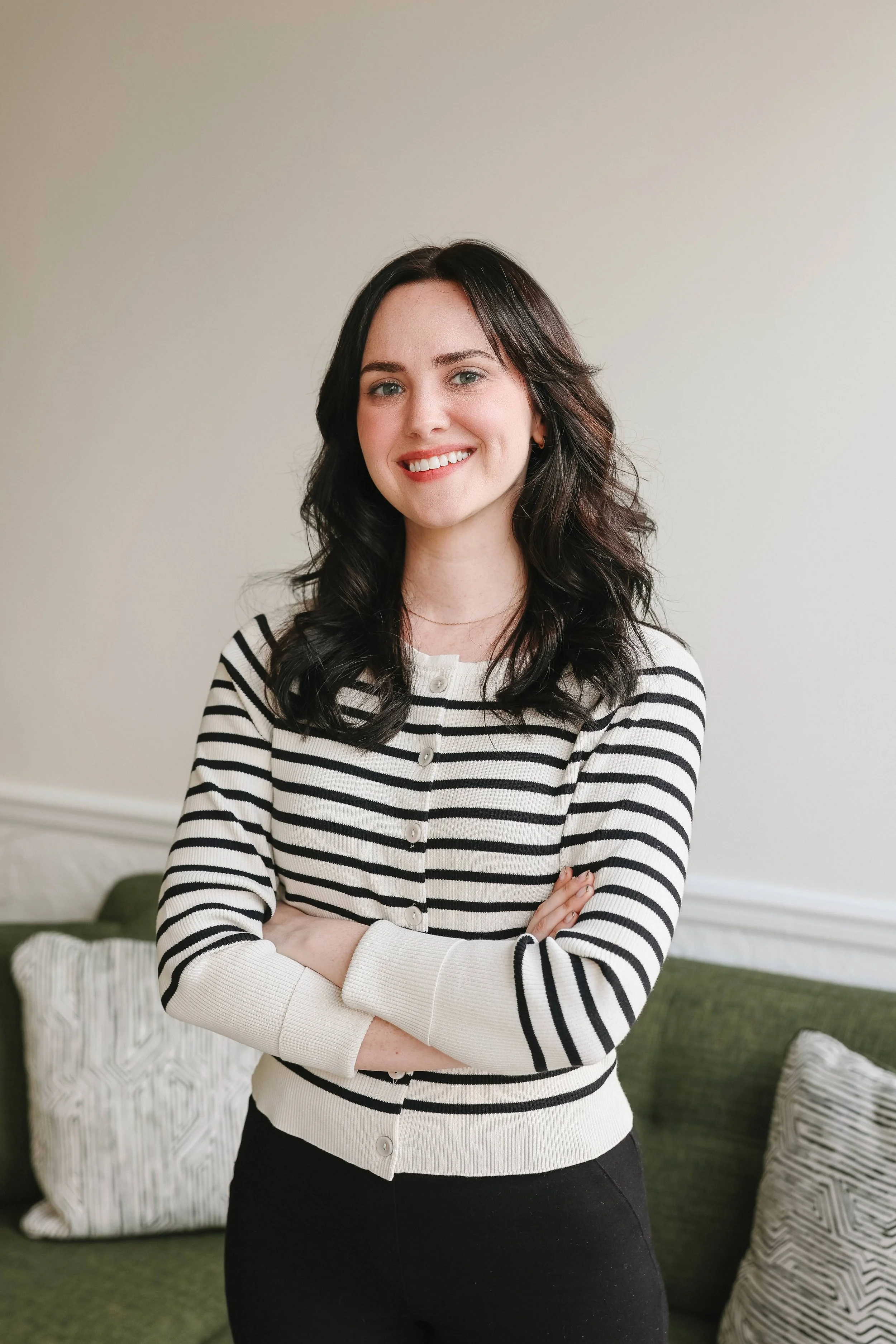A woman with dark, wavy hair smiling with her arms crossed, wearing a black and white striped cardigan, standing in a room with a green couch and patterned pillows.