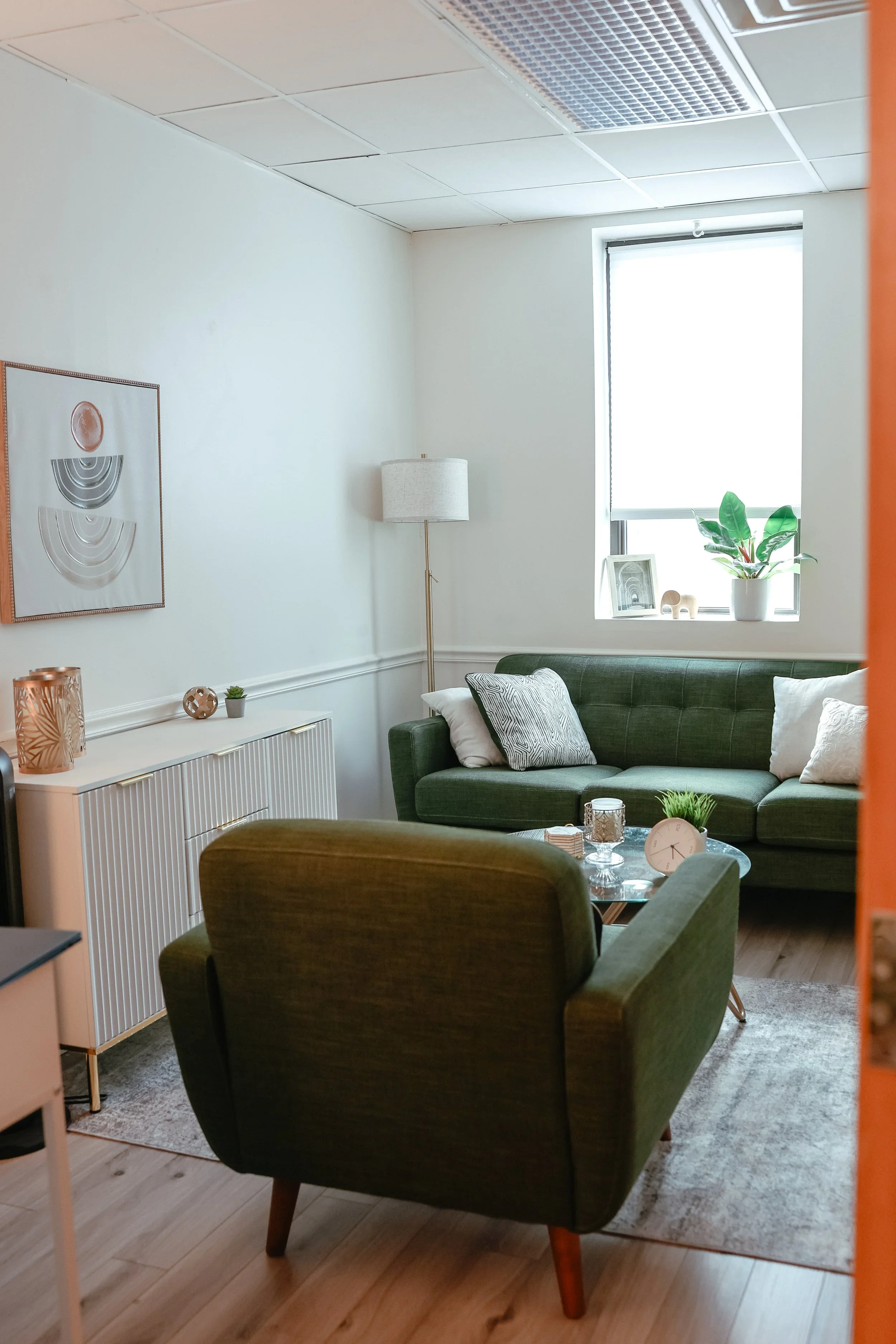 A cozy living room with a green sofa and armchair, a coffee table with decorative items, a window with natural light, indoor plants, artwork on the wall, and a lamp.