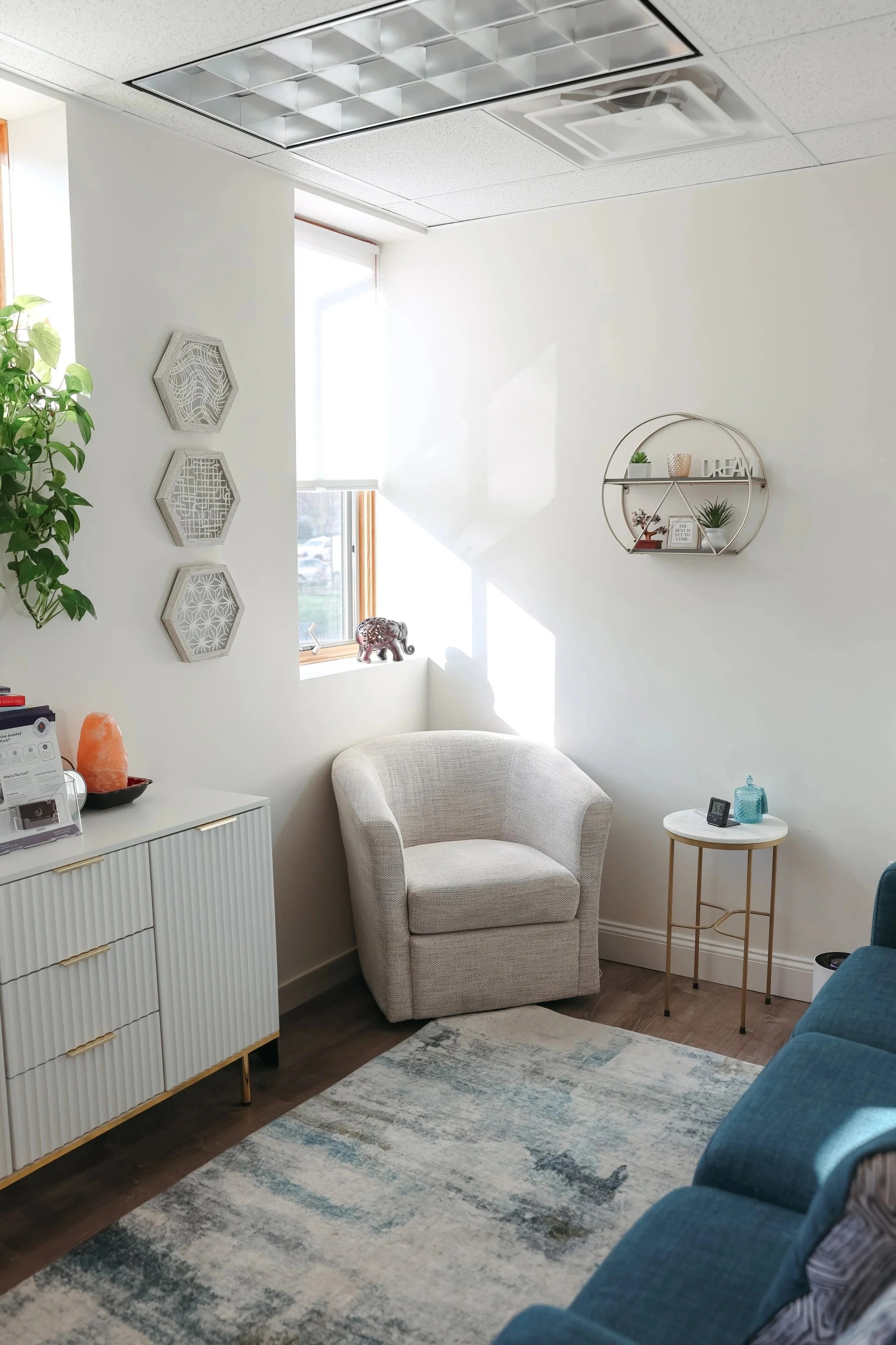 A cozy corner of a living room with a white armchair, a small round side table with decor items, a turquoise couch partially visible, a white cabinet with a salt lamp on top, decorative wall art, a window letting in natural light, and a patterned area rug.