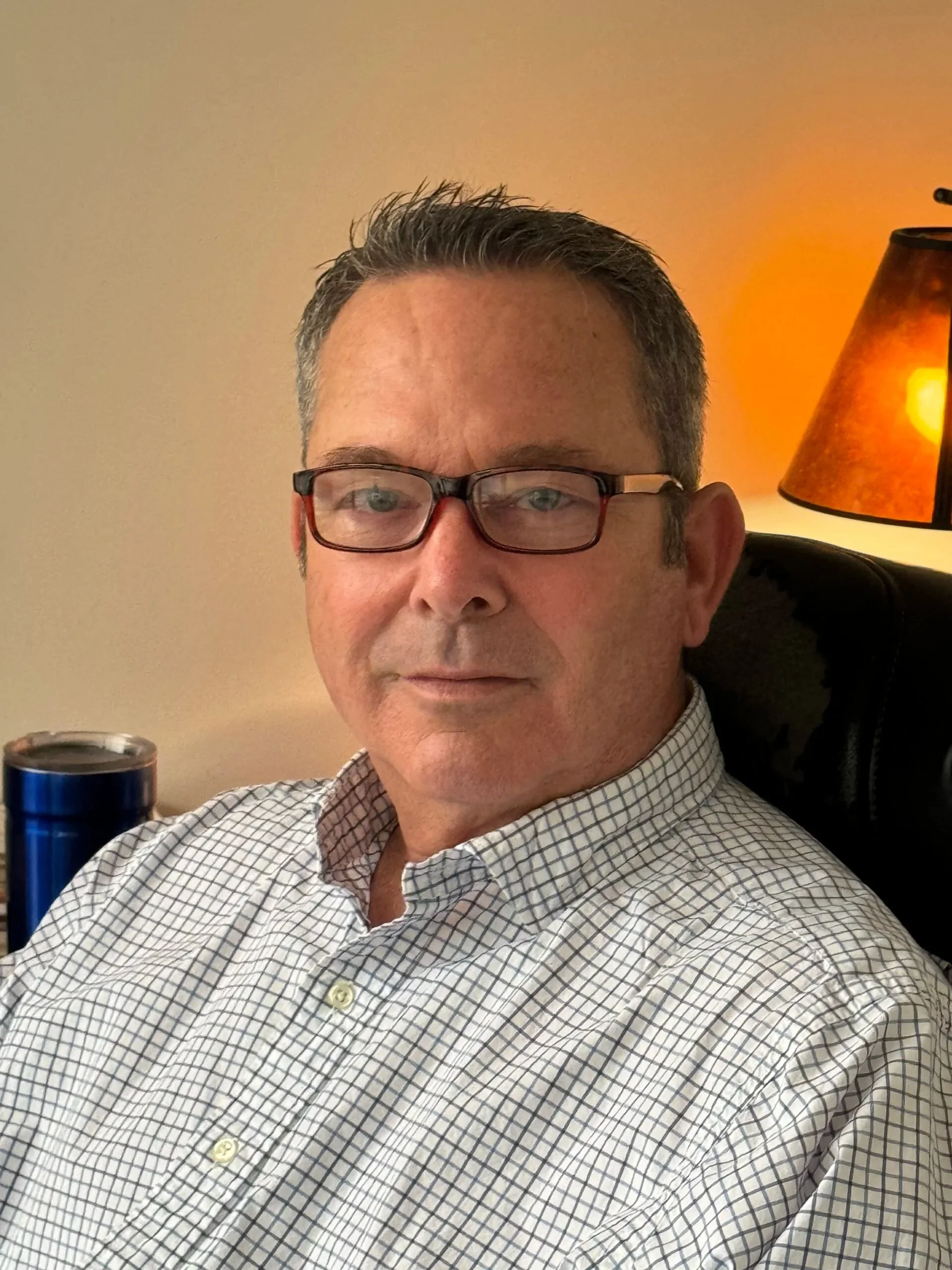 A middle-aged man with short dark hair, glasses, and a checkered shirt sitting in a black chair with a brown lamp and beige wall in the background.