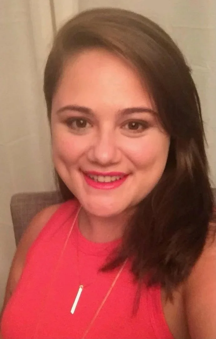 A young woman with brown hair, wearing a pink sleeveless top and a white necklace, smiling at the camera.