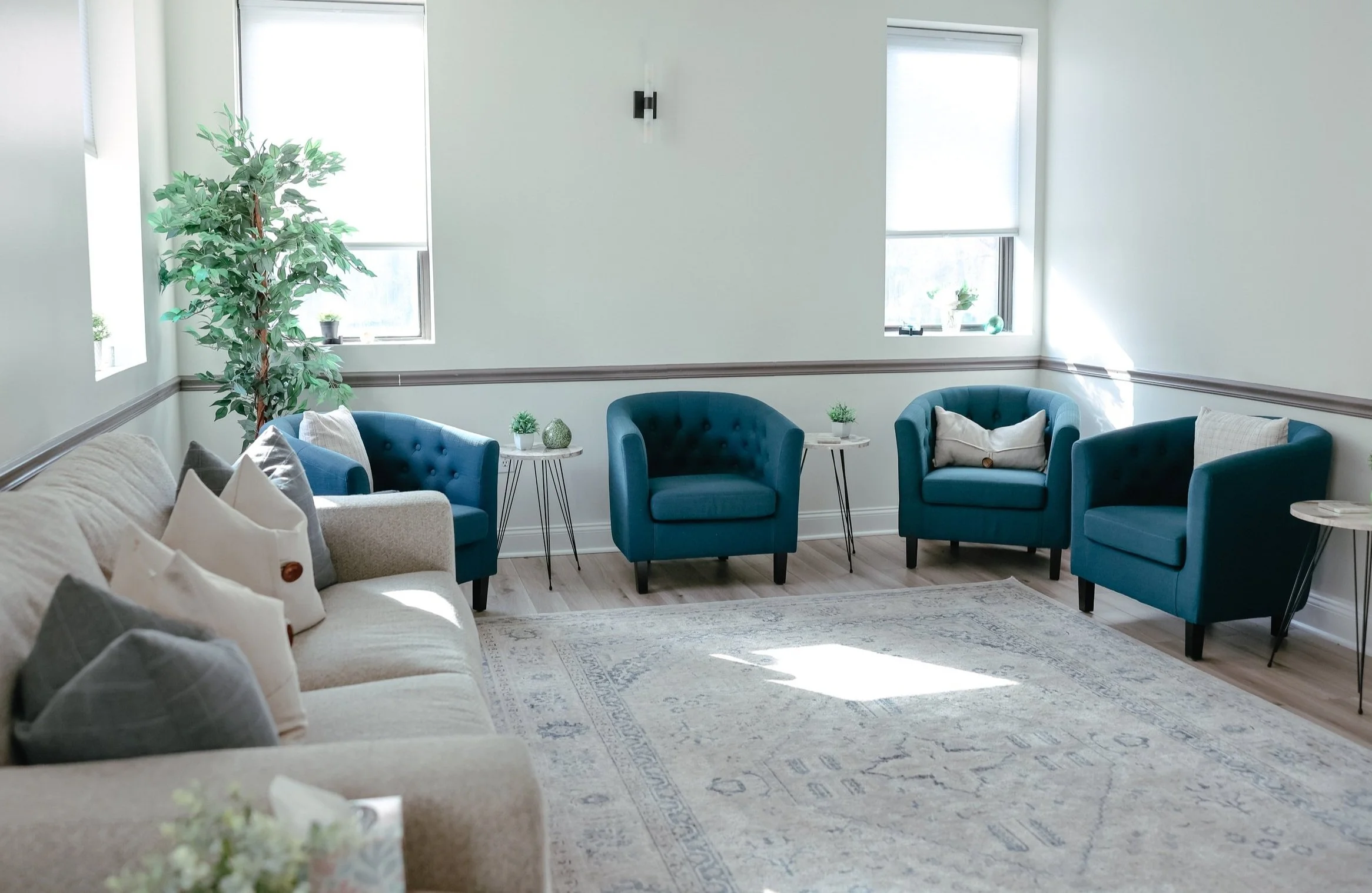 A bright living room with beige sofa and three teal armchairs arranged in a semi-circle, small white side tables with decorative items, large windows with white blinds, a large indoor plant, and a light-colored area rug.
