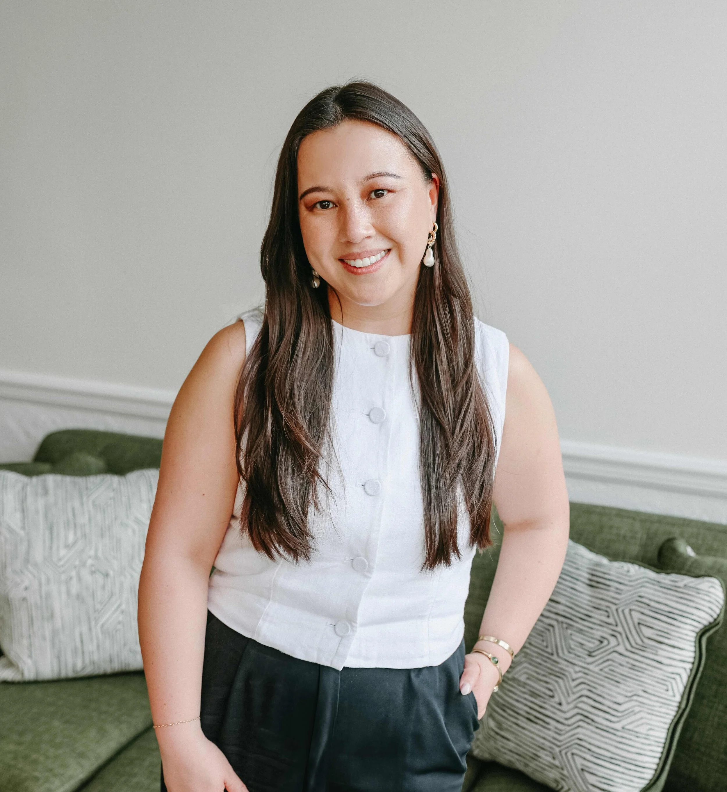 A woman with long dark hair, wearing a white sleeveless button-up top and black pants, smiling and standing in front of a green couch with patterned cushions.