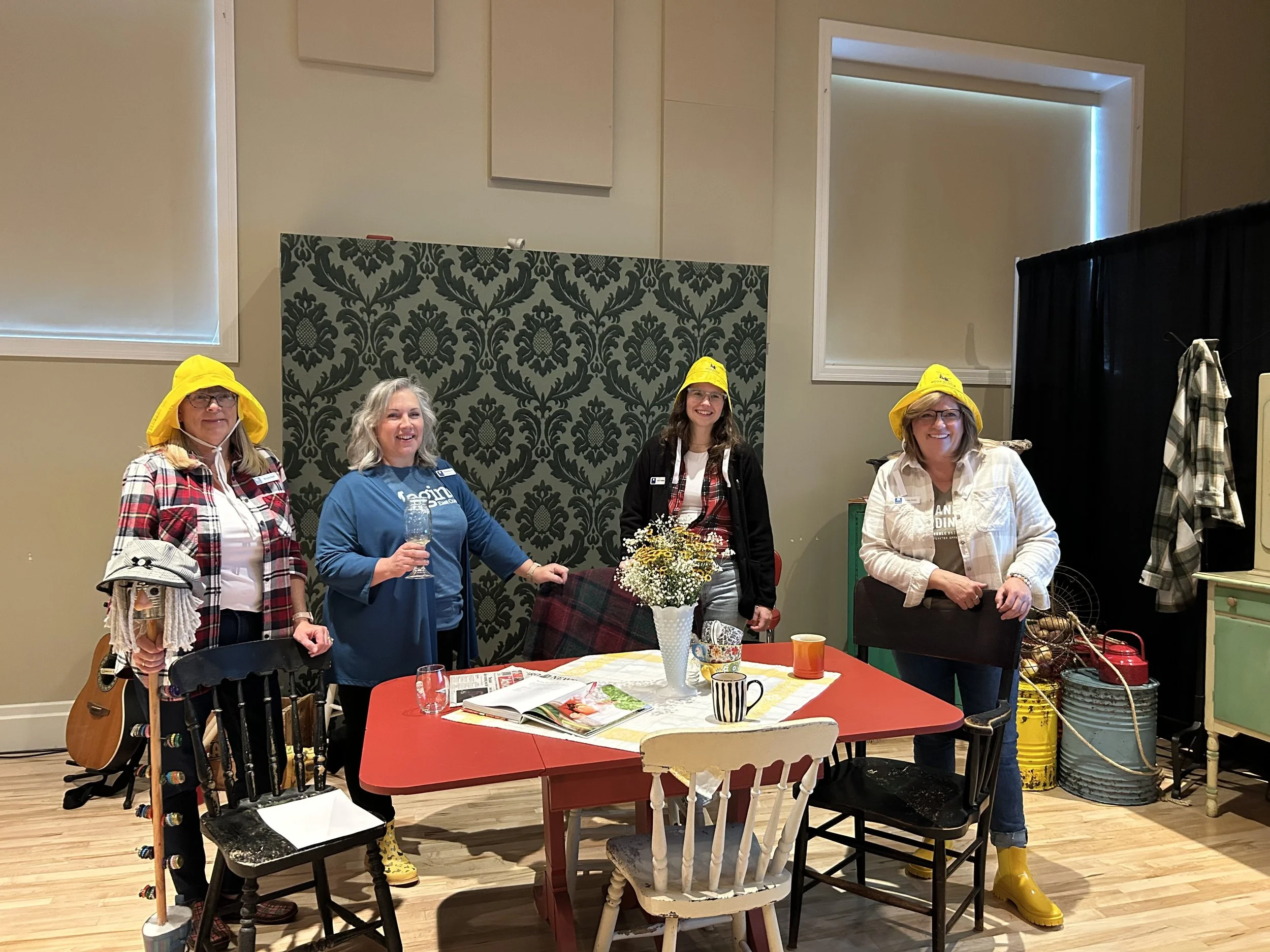 Four women standing around a dining table with a flower vase and cups, wearing yellow rain hats and casual clothes, in a room with green patterned wallpaper and black curtains.