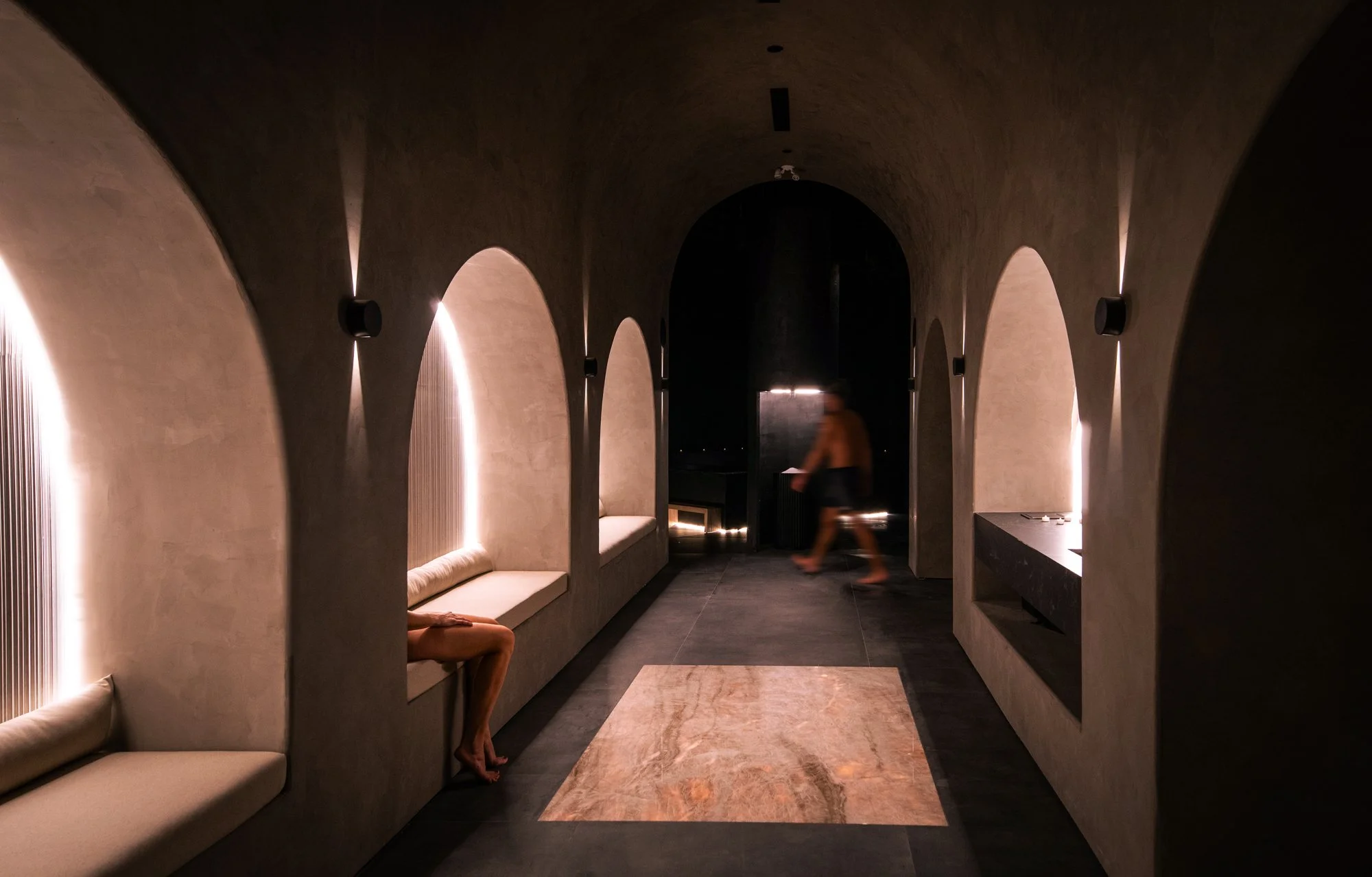 Dimly lit corridor with arched alcoves having seating, a person walking in the background, and a beige marble tile on the floor.
