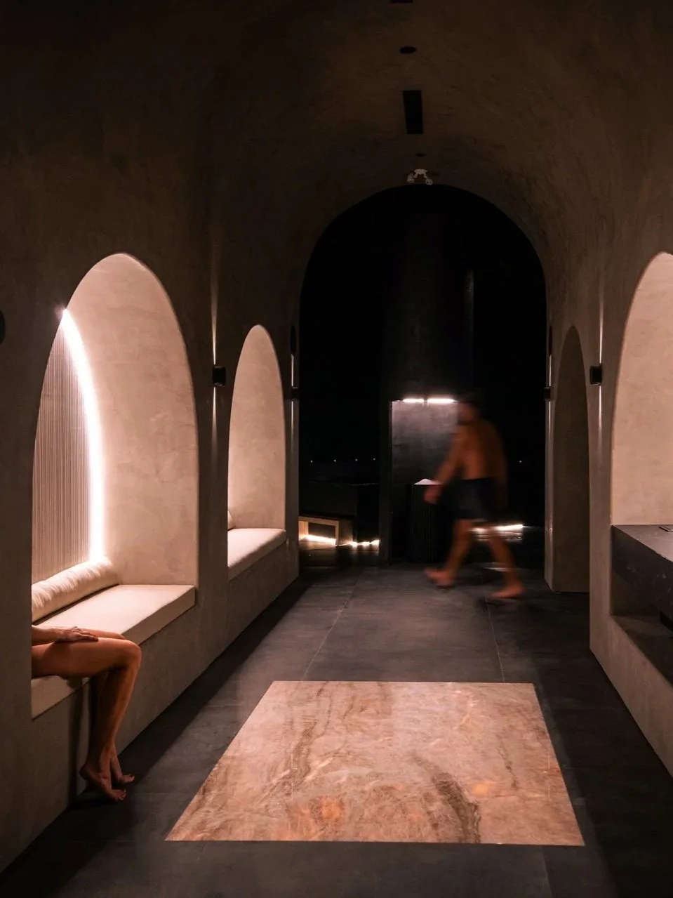 Interior view of a spa featuring a rounded ceiling, recessed seating nooks, and steam flowing from a sauna door, with a moody, cozy atmosphere and natural materials like clay paint and onyx stone.