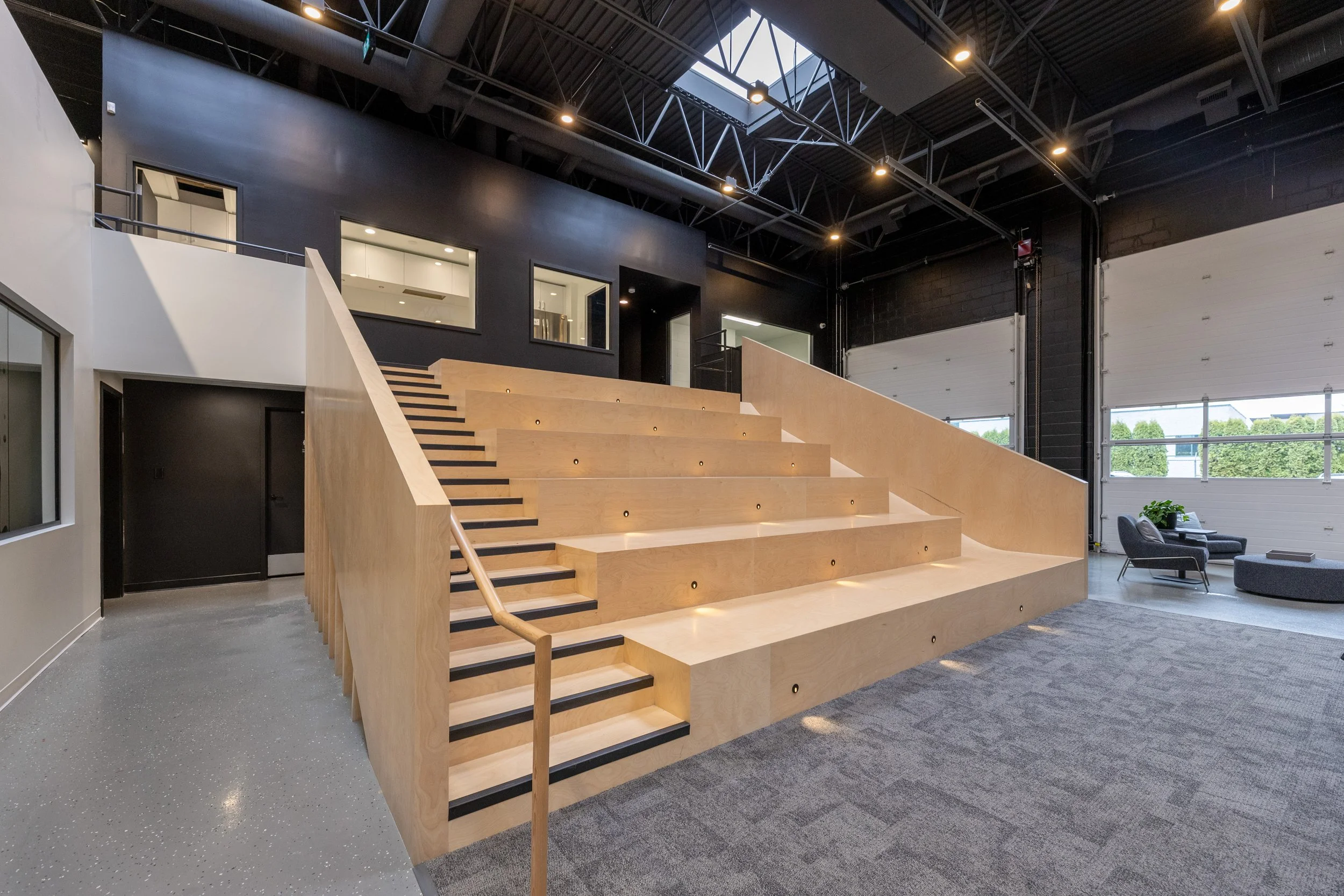 Office lounge with black and wood accents, featuring a seating area, bar with four chairs, leafy plants in a built-in planter, large glass windows, and a black curving staircase leading to the upper level.