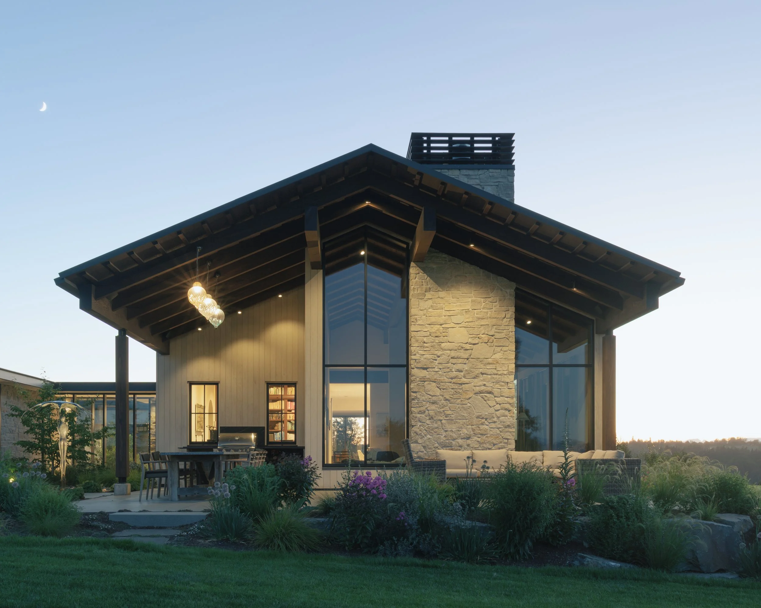 Interior view of a modern living room with high vaulted wooden ceiling, large windows overlooking the outdoors, a stone fireplace, and comfortable seating furniture. The outside scene shows mountains and trees.