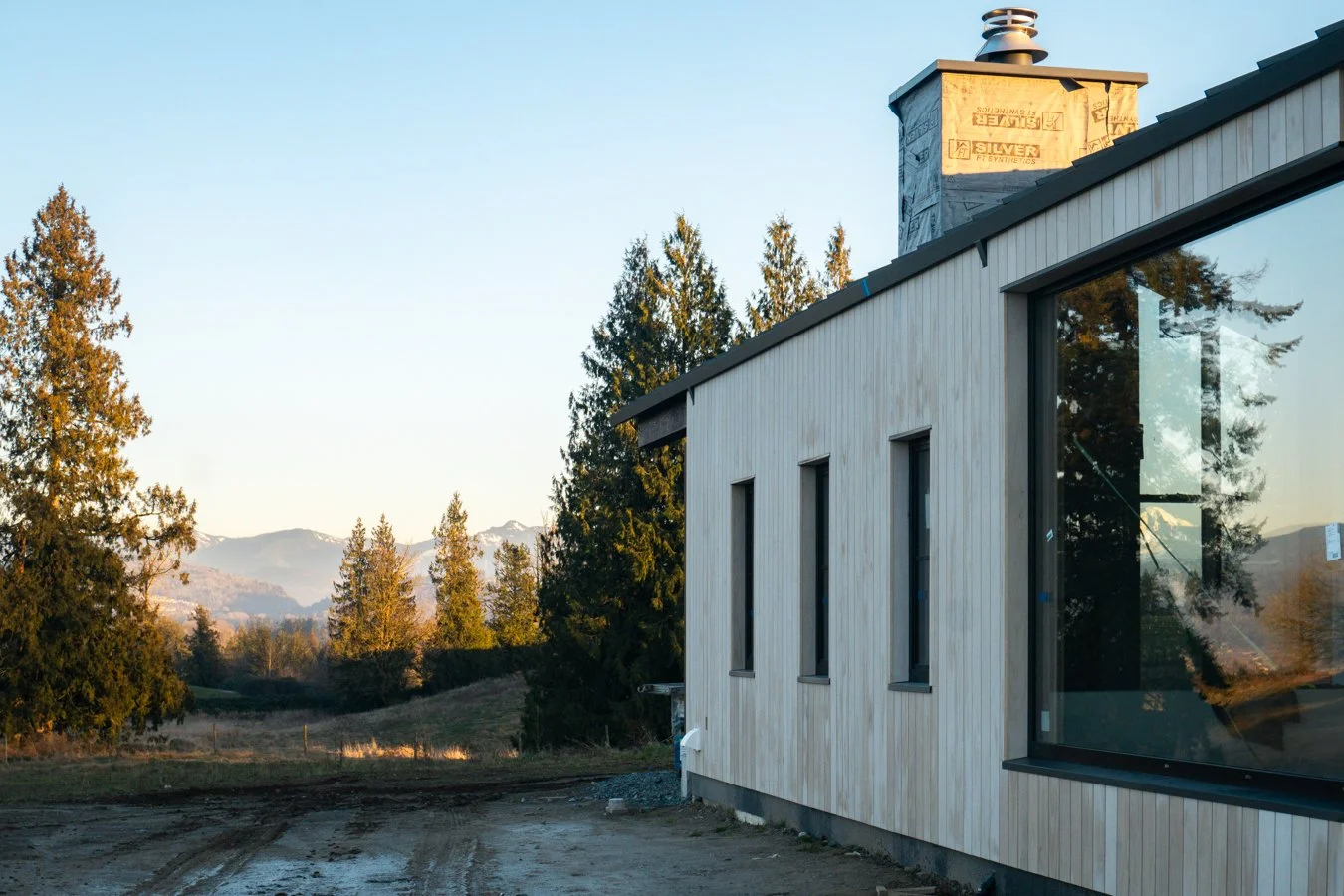 A modern house with wooden siding and large windows, situated in a rural area with trees and mountains in the background, during sunset.