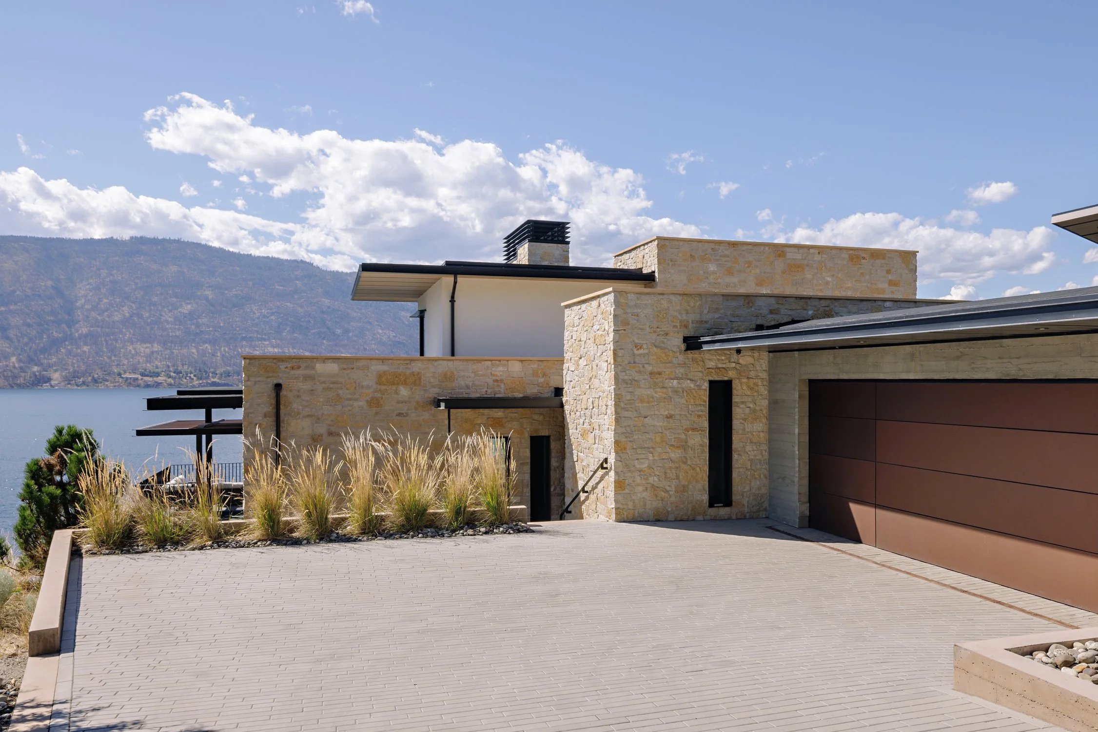 Modern house with a stone facade, large garage door, and a view of a lake and mountains in the background on a sunny day.