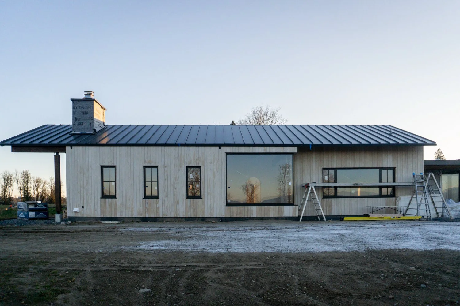Under construction modern house with light wood exterior, black-framed windows, metal roof, and a chimney, set against a clear sky.