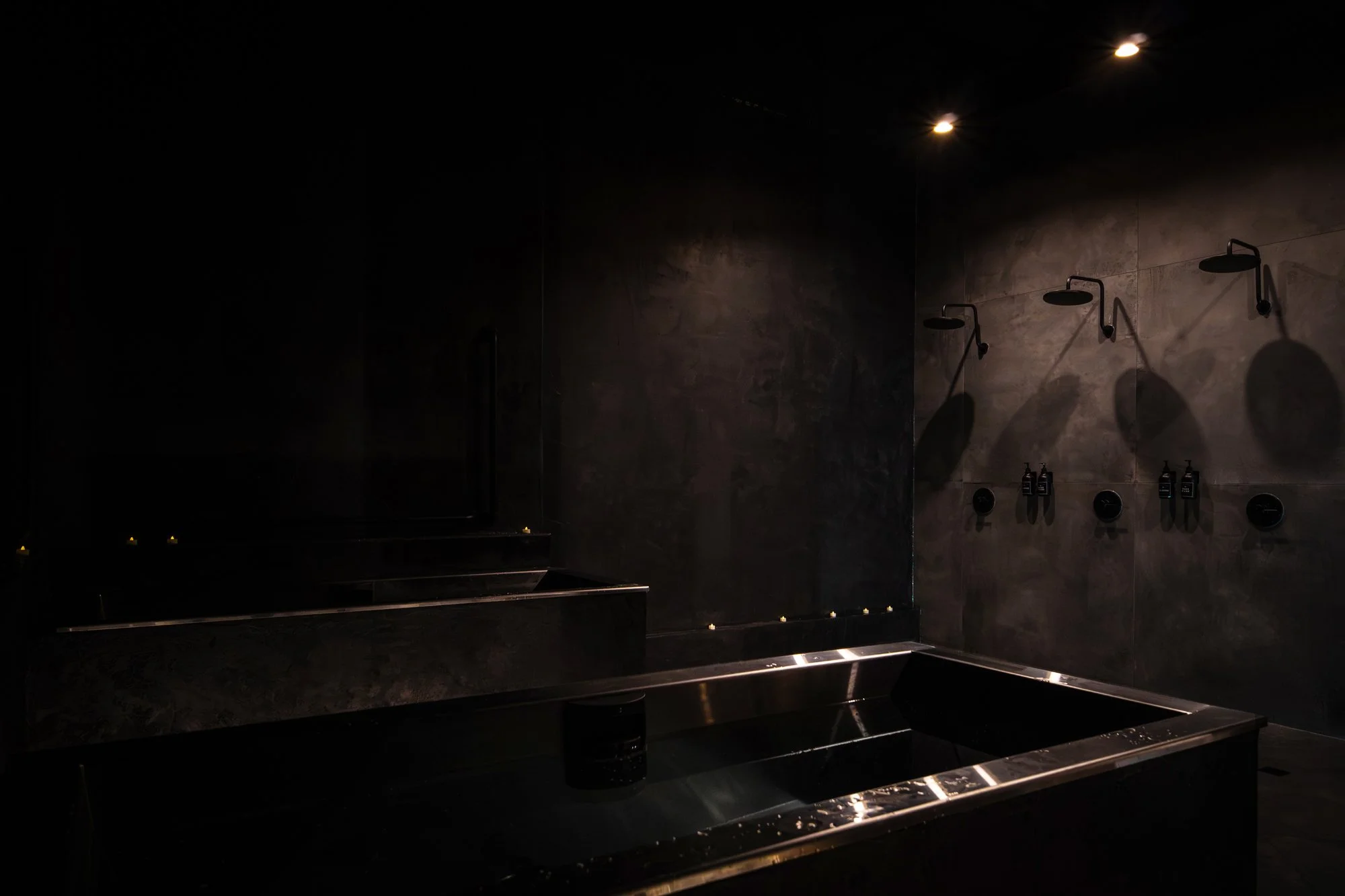 Dark, minimalist shower room with three overhead showerheads on a gray tiled wall, a built-in bathtub, and dim lighting.