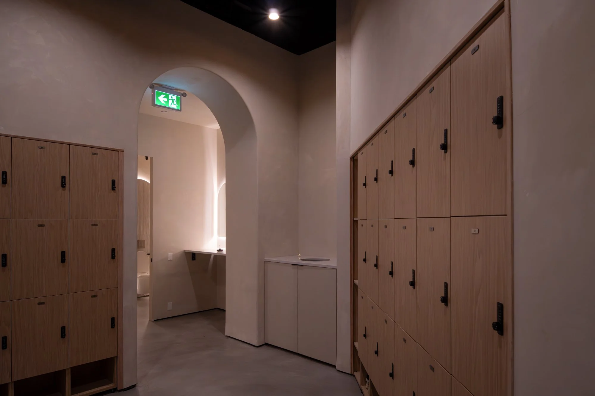 A hallway with wooden lockers on both sides, an arched doorway at the end with a green emergency exit sign, and a small sink with a cabinet underneath and a mirror on the wall beyond the opening.