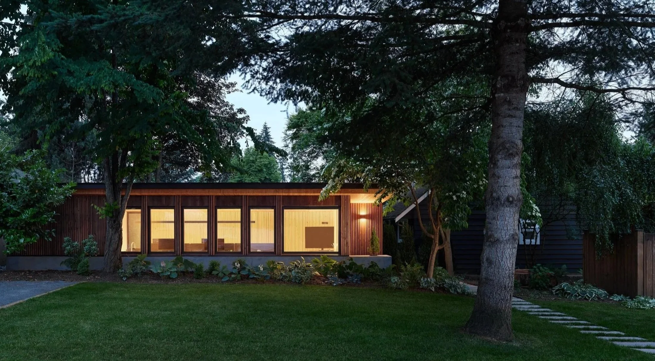 Modern house with large windows, wooden exterior, surrounded by trees and a well-maintained lawn, with outdoor lighting on.
