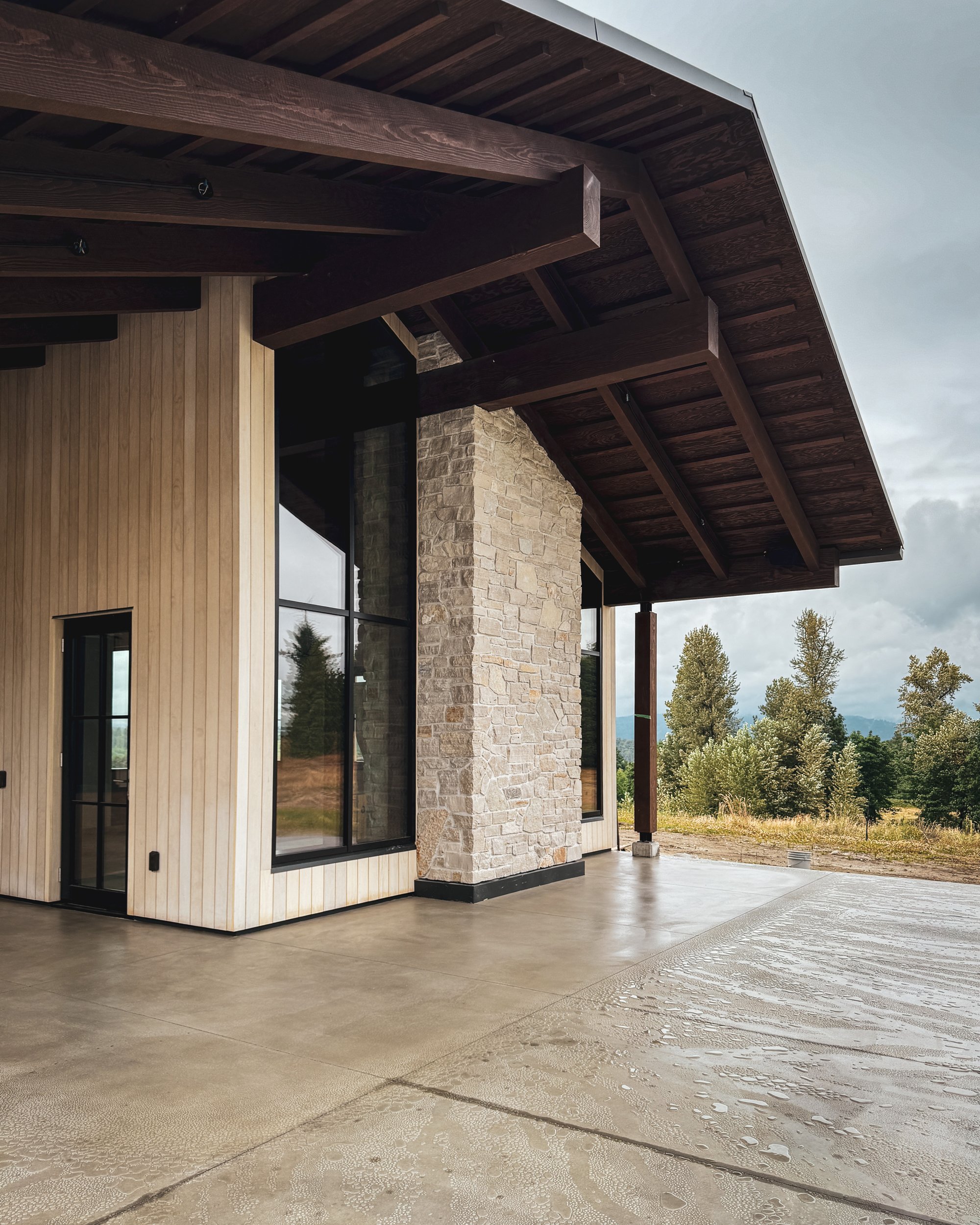 Single-story contemporary home surrounded by landscaping, made of natural materials like stone and wood, featuring a gable roof and a swimming pool in the front yard.