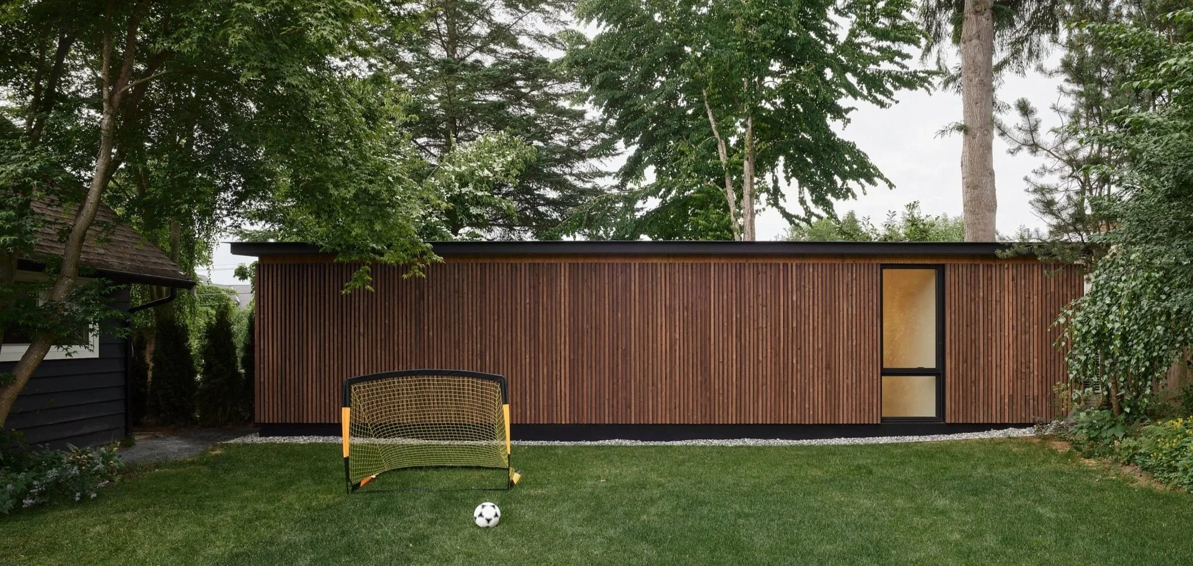 Backyard with green grass, a small soccer goal, and a soccer ball. There's a modern wooden shed or tiny house in the background surrounded by trees.