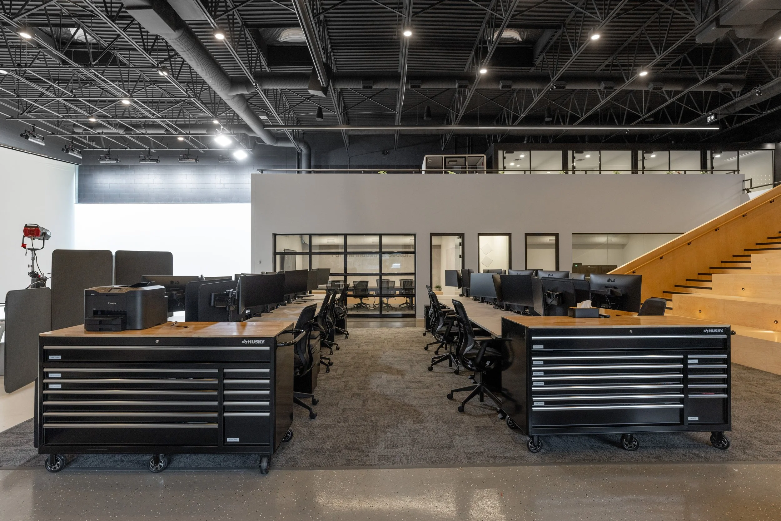 Open-plan office featuring multiple workstations, a riser seating area leading to an upper level, a lounge space, and glass-walled meeting rooms.
