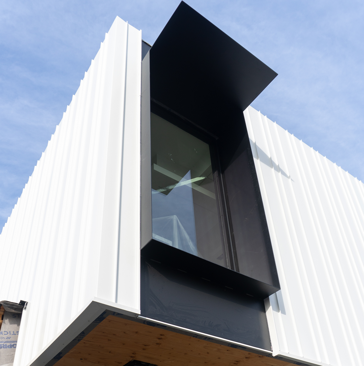 Modern building exterior with white vertical siding, large black-framed window, and a black overhang, against a blue sky.