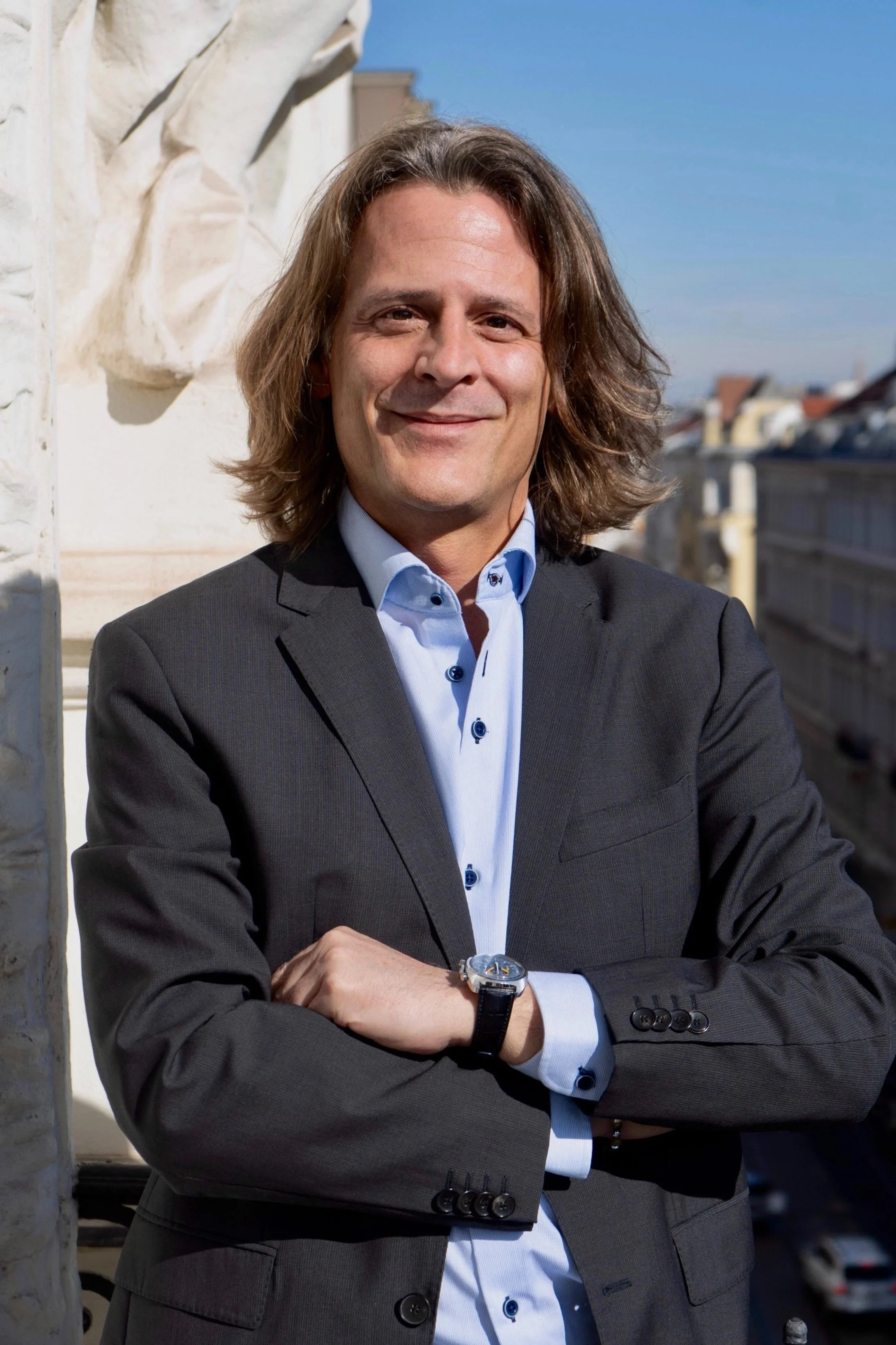 Ein Mann in Anzug mit verschränkten Armen auf einem Balkon, im Hintergrund Stadtansicht, sonniger Tag.